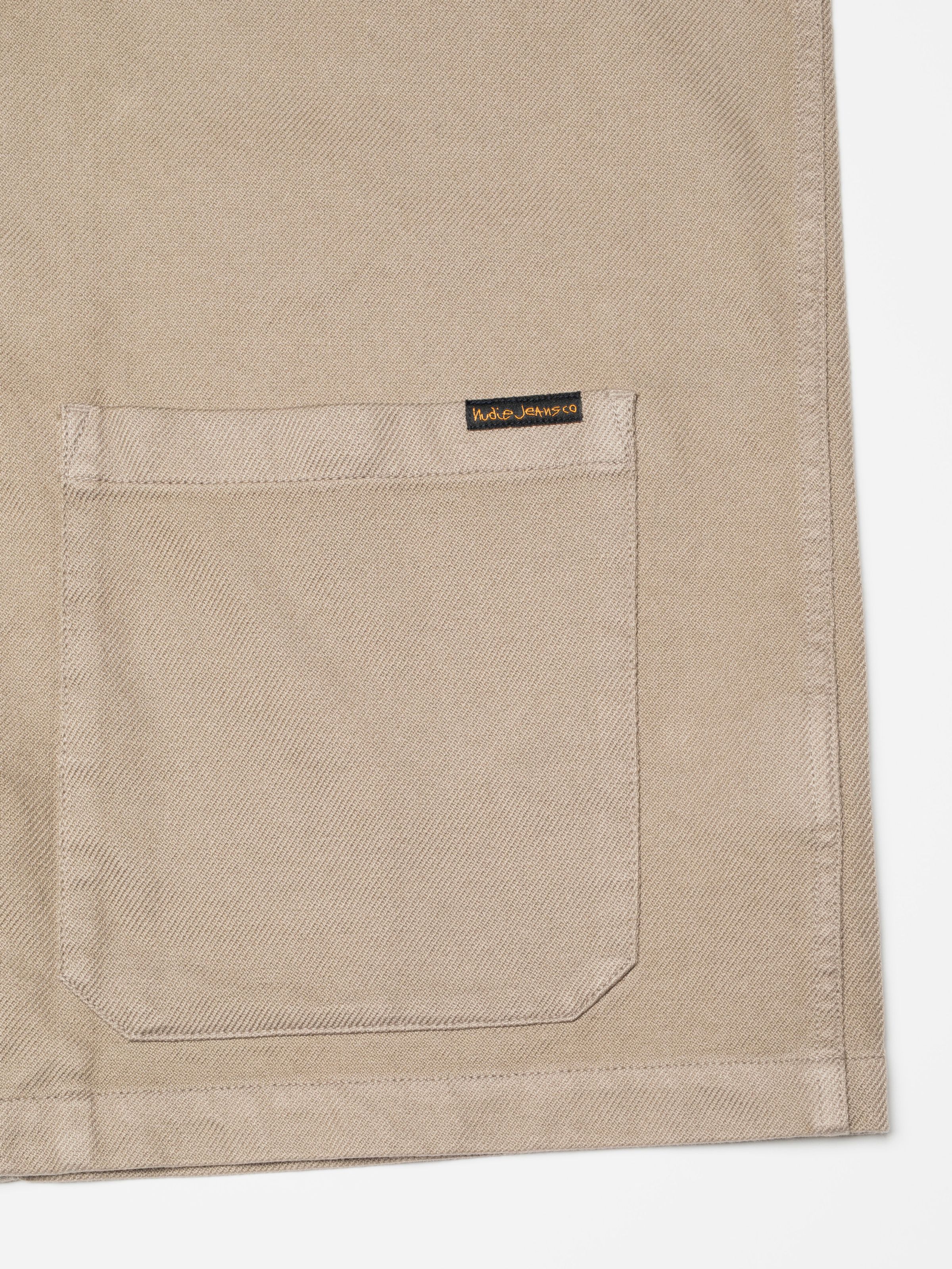 Nudie Jeans Barney Worker Jacket in Mud, close-up of the patch pocket and black logo label.