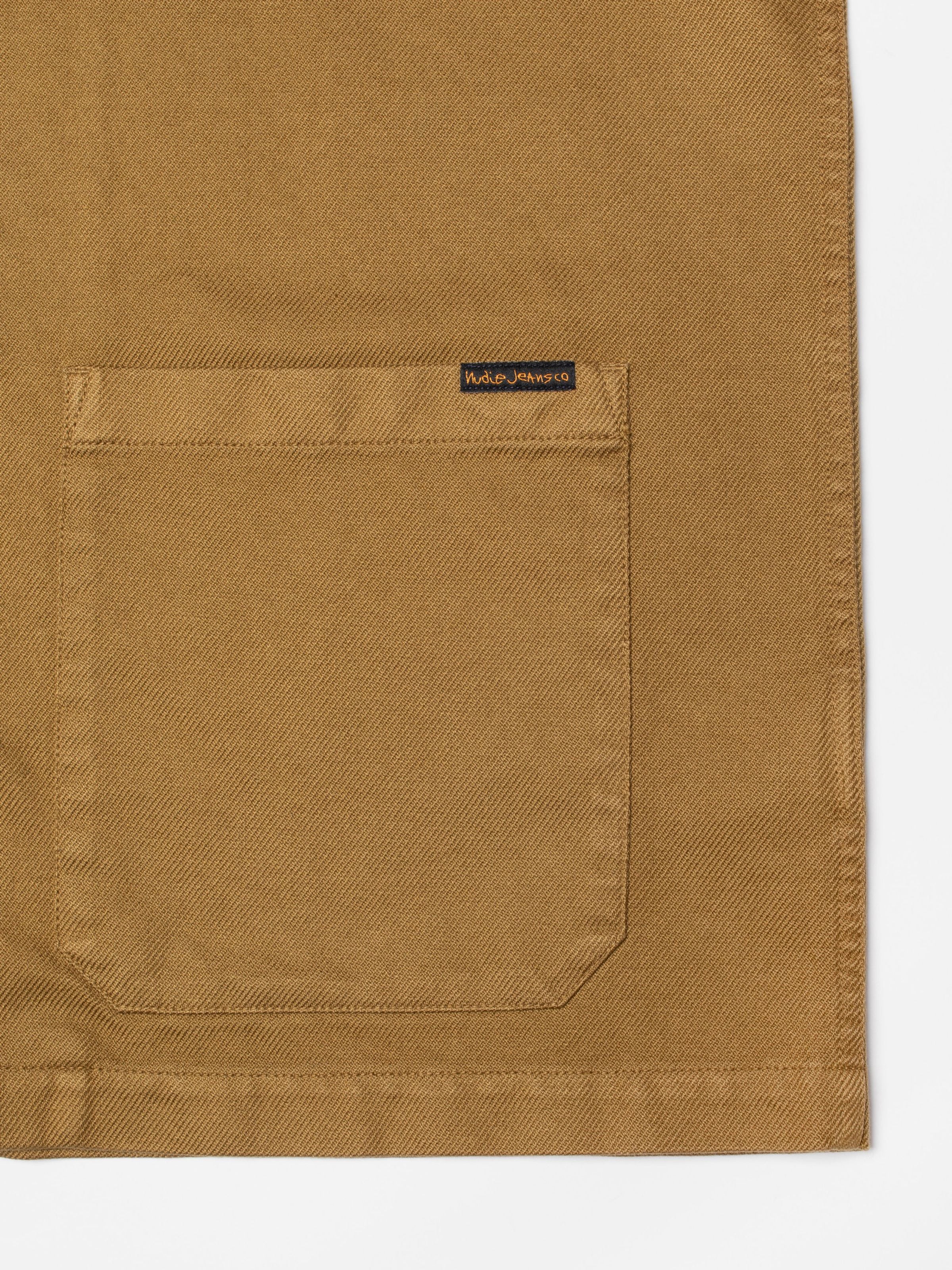Nudie Jeans Barney Worker Jacket in Braun-Olive, Nahaufnahme der aufgesetzten Vordertasche und der Twill-Stoffstruktur.