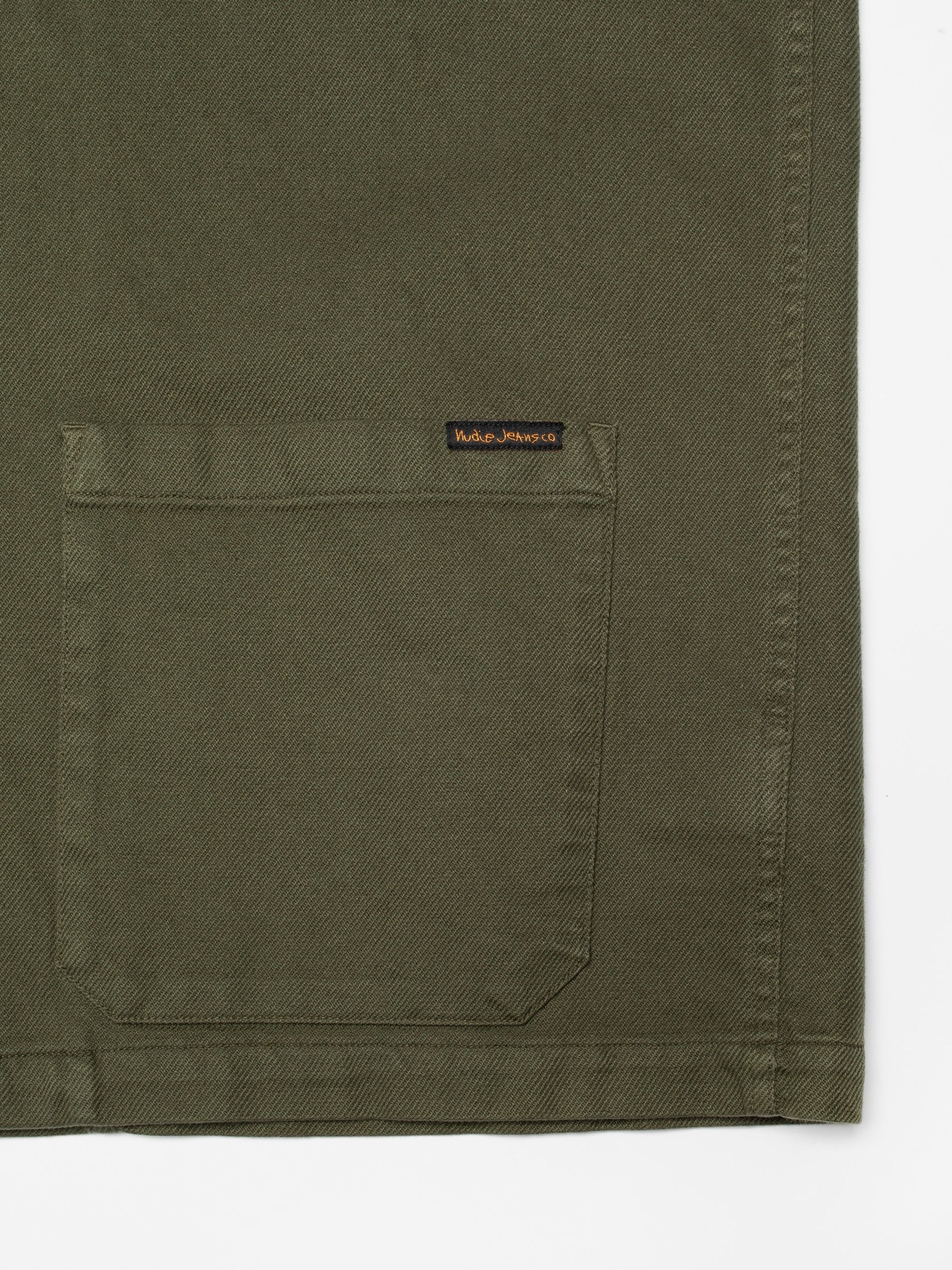 Nudie Jeans Barney Worker Jacket Olive, close-up of a patch pocket showing the small Nudie Jeans logo label.
