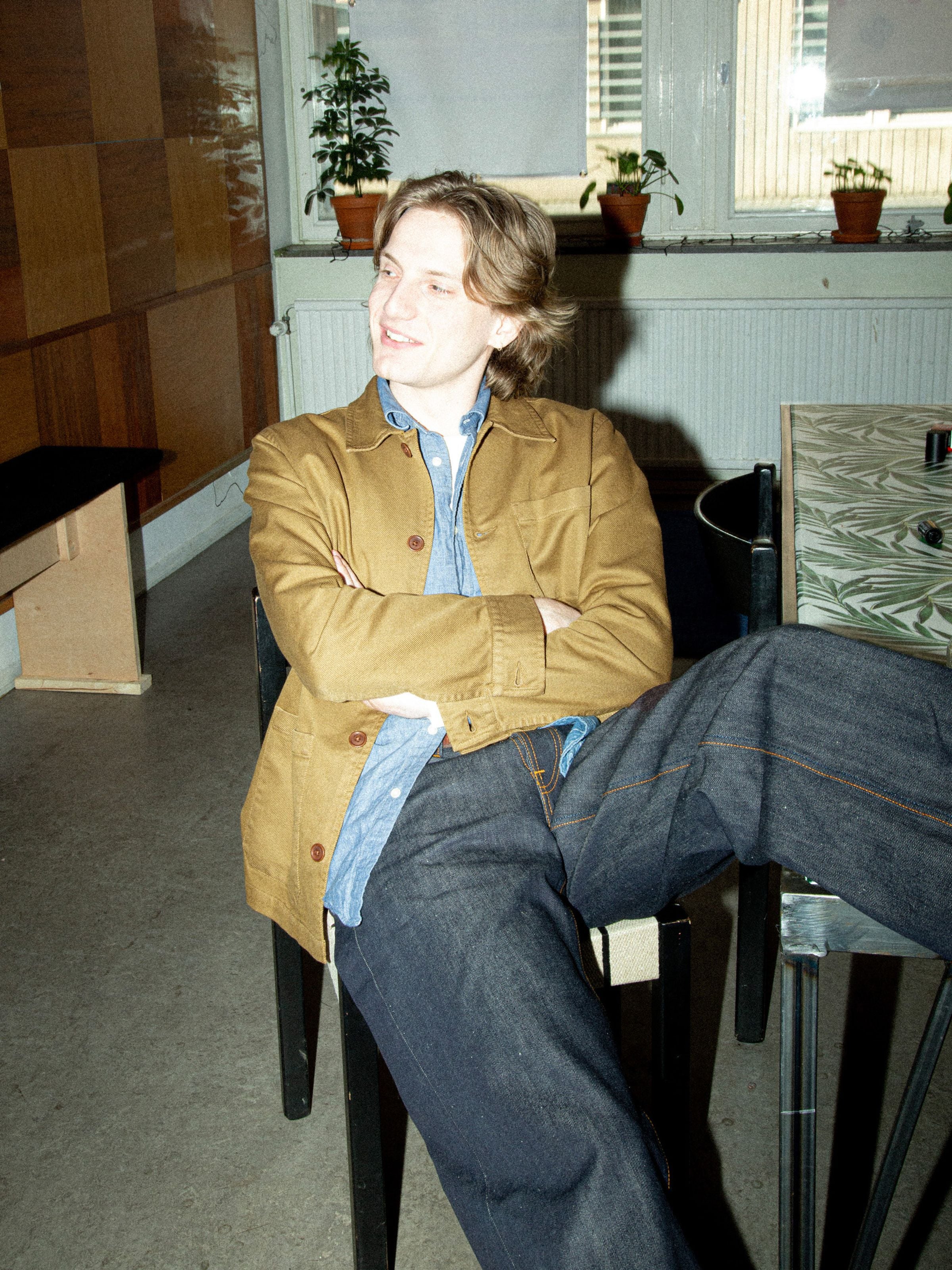 Nudie Jeans Barney Worker Jacket in Braun-Olive von einem Mann getragen, der im Innenbereich sitzt.