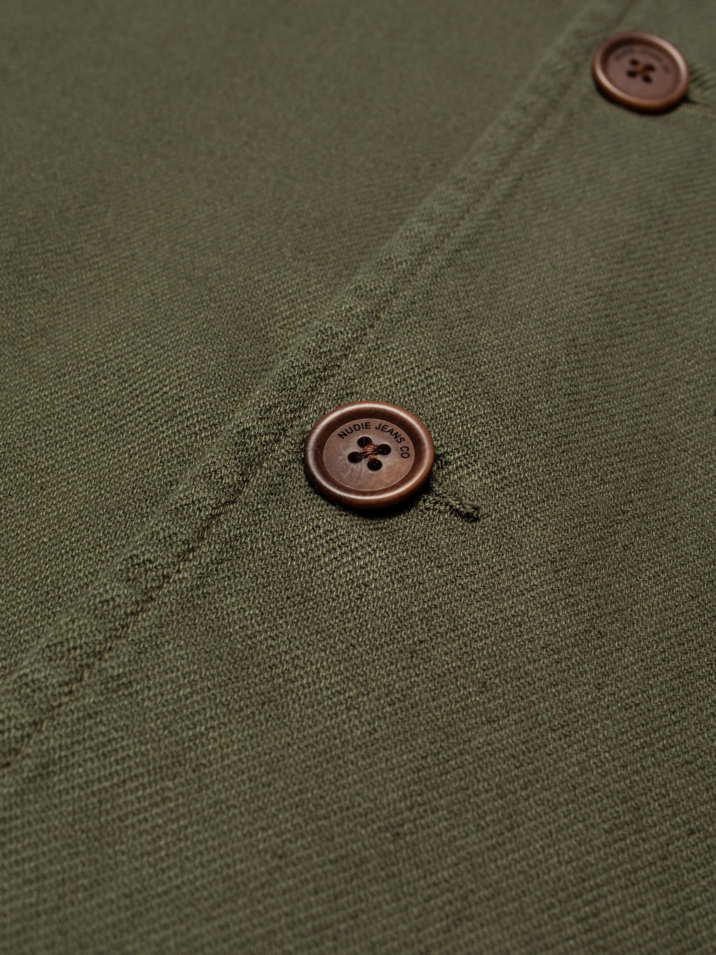 Nudie Jeans Barney Worker Jacket in Olive, a close-up shot of the twill fabric and logo-engraved button.