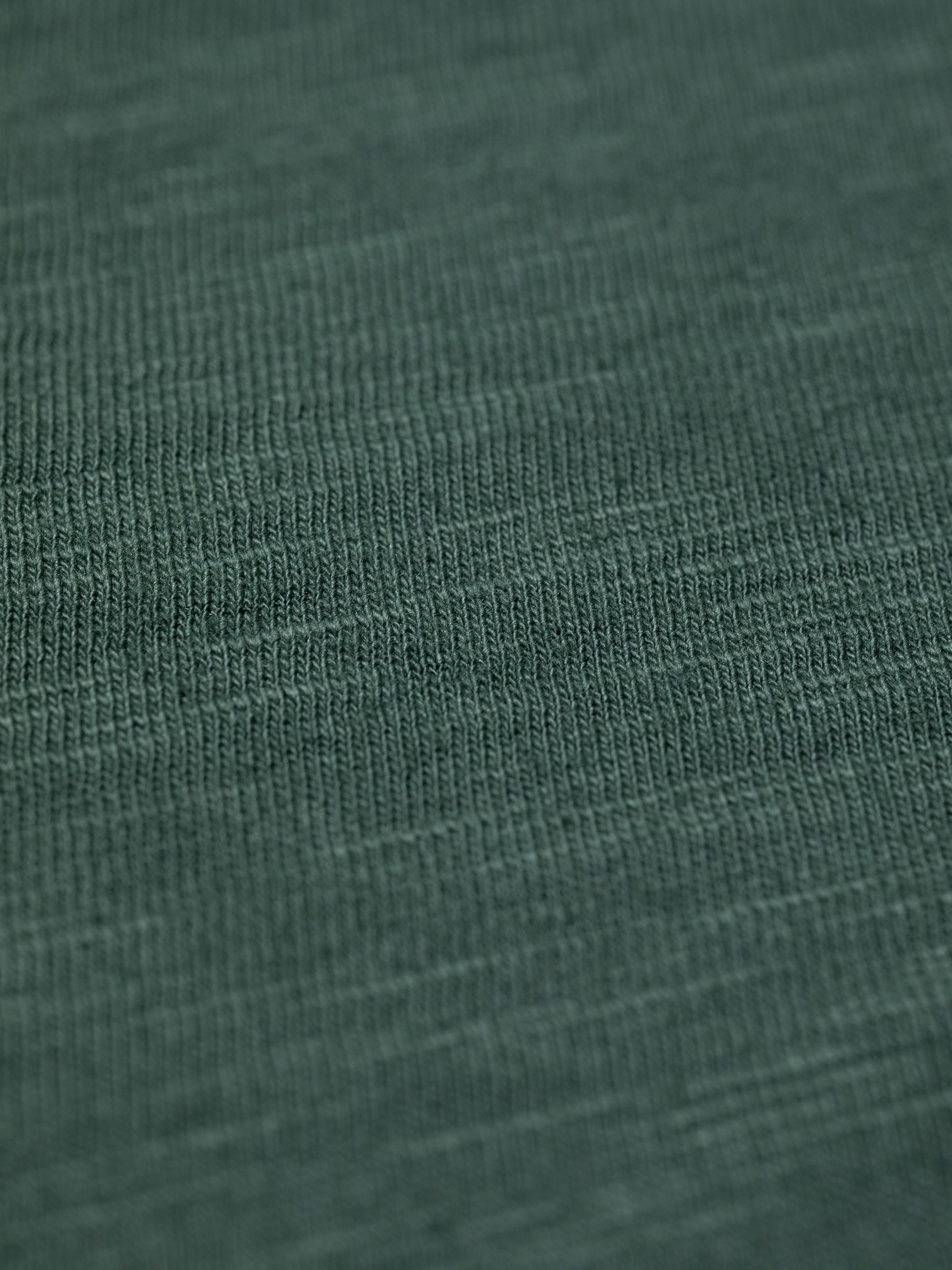 Nudie Jeans Roffe Slub T-Shirt in Dunkelgrün, Nahaufnahme des Bio-Baumwolljerseys mit Garnverdickungen.