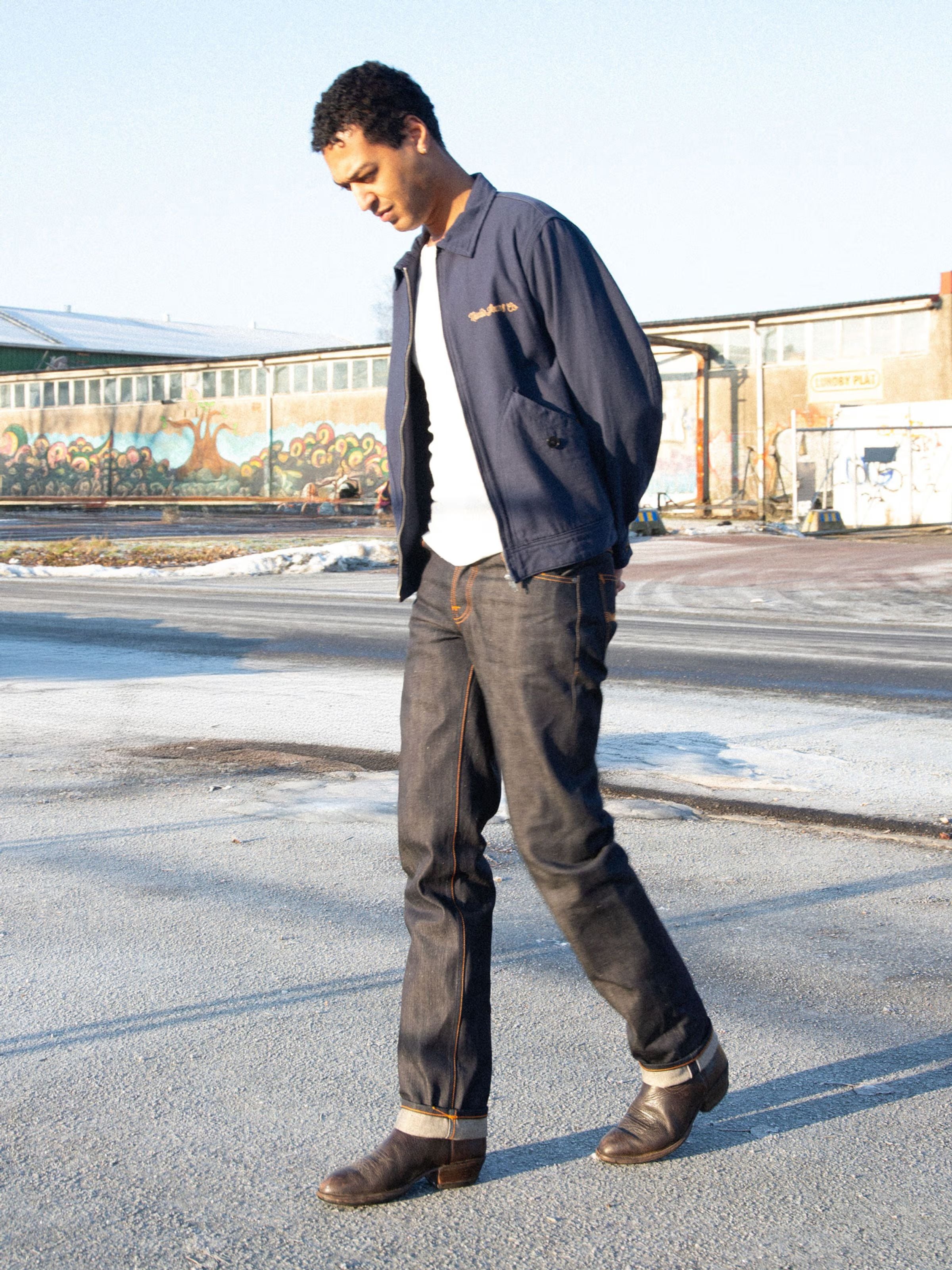 Nudie Jeans Rad Rufus Dry Selvage, men's jeans in dry selvedge denim, worn outdoors against an industrial backdrop.