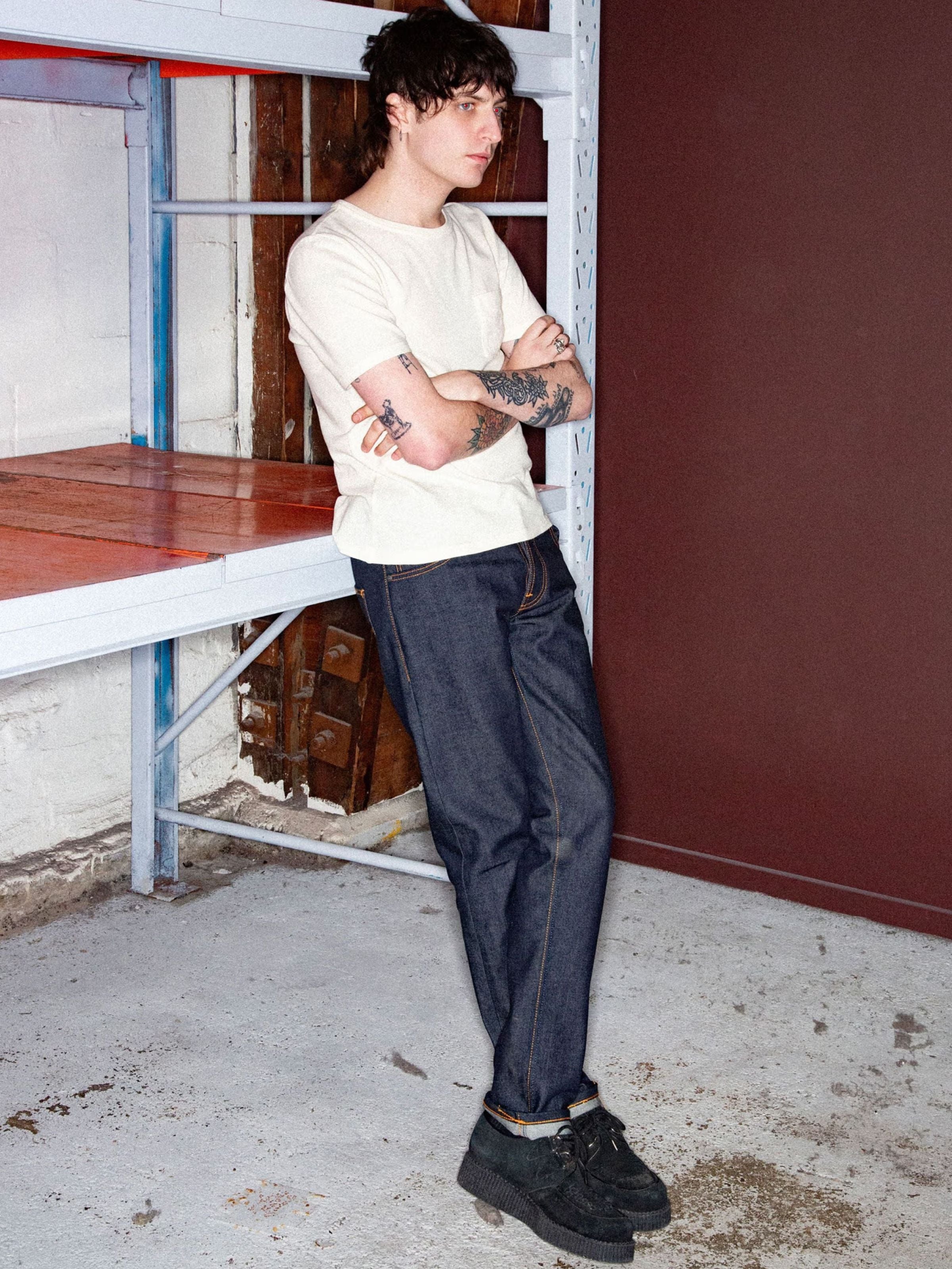 Nudie Jeans Grim Tim Dry Japan Selvage, men's slim selvedge jeans in dark indigo, lifestyle shot leaning against a rack.