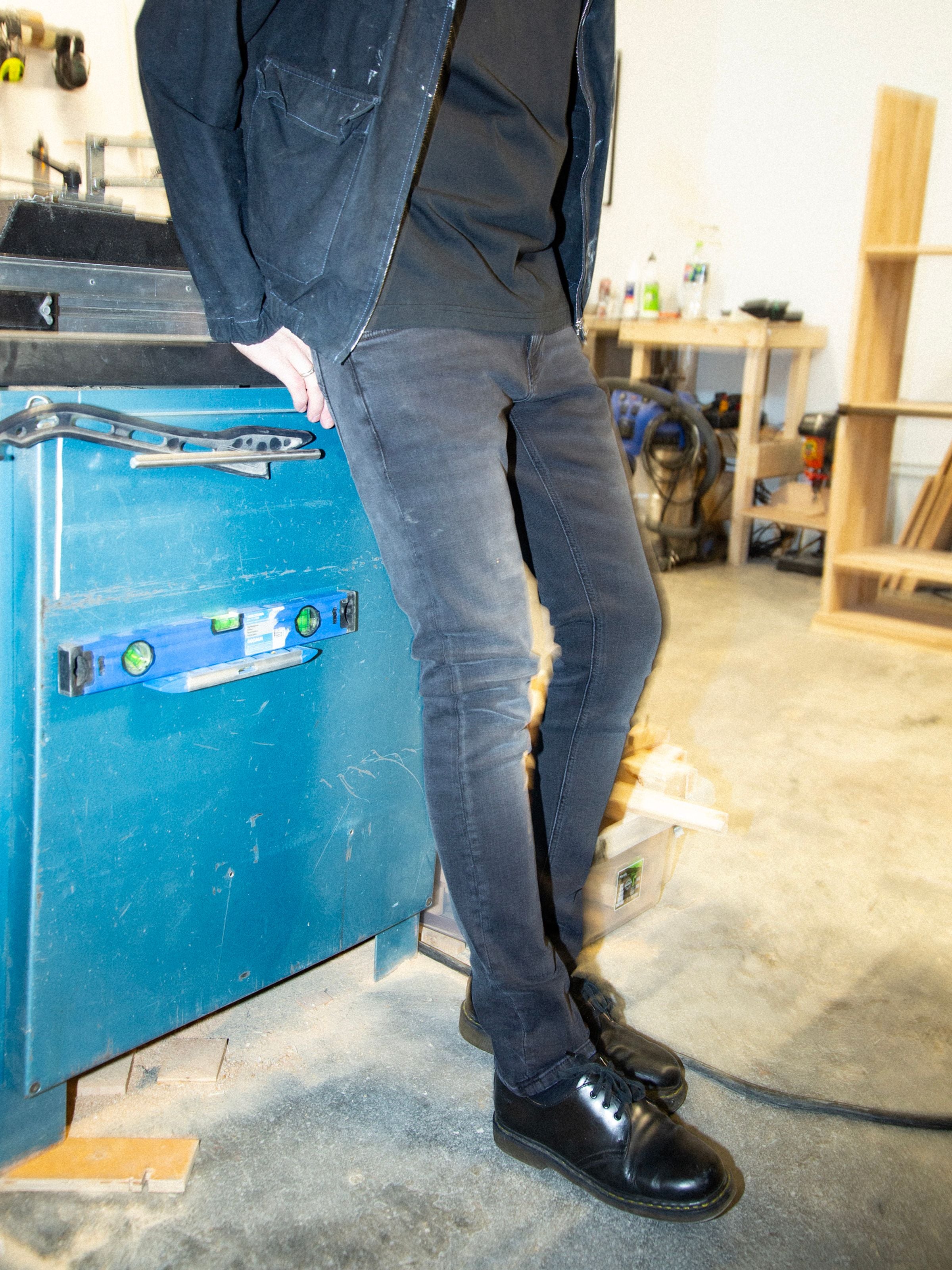 Nudie Jeans Tight Terry Black Blizzard, Herrenjeans mit enger Passform, von der Taille abwärts in einer Werkstatt gezeigt.