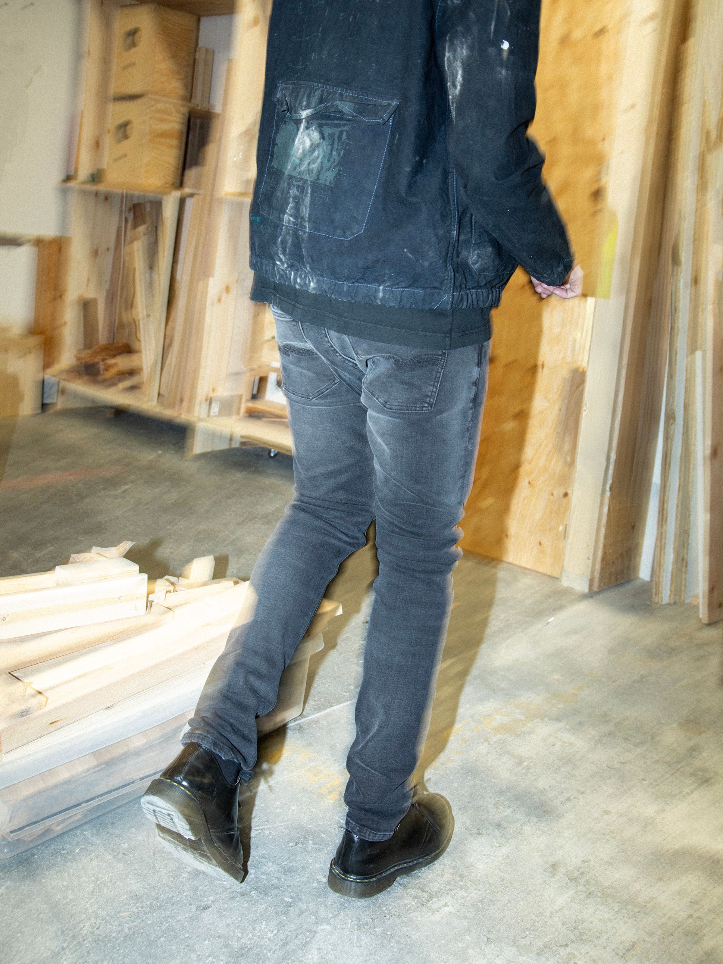 Nudie Jeans Tight Terry Black Blizzard, men's tight fit jeans, shown from the back on a model in a workshop setting.