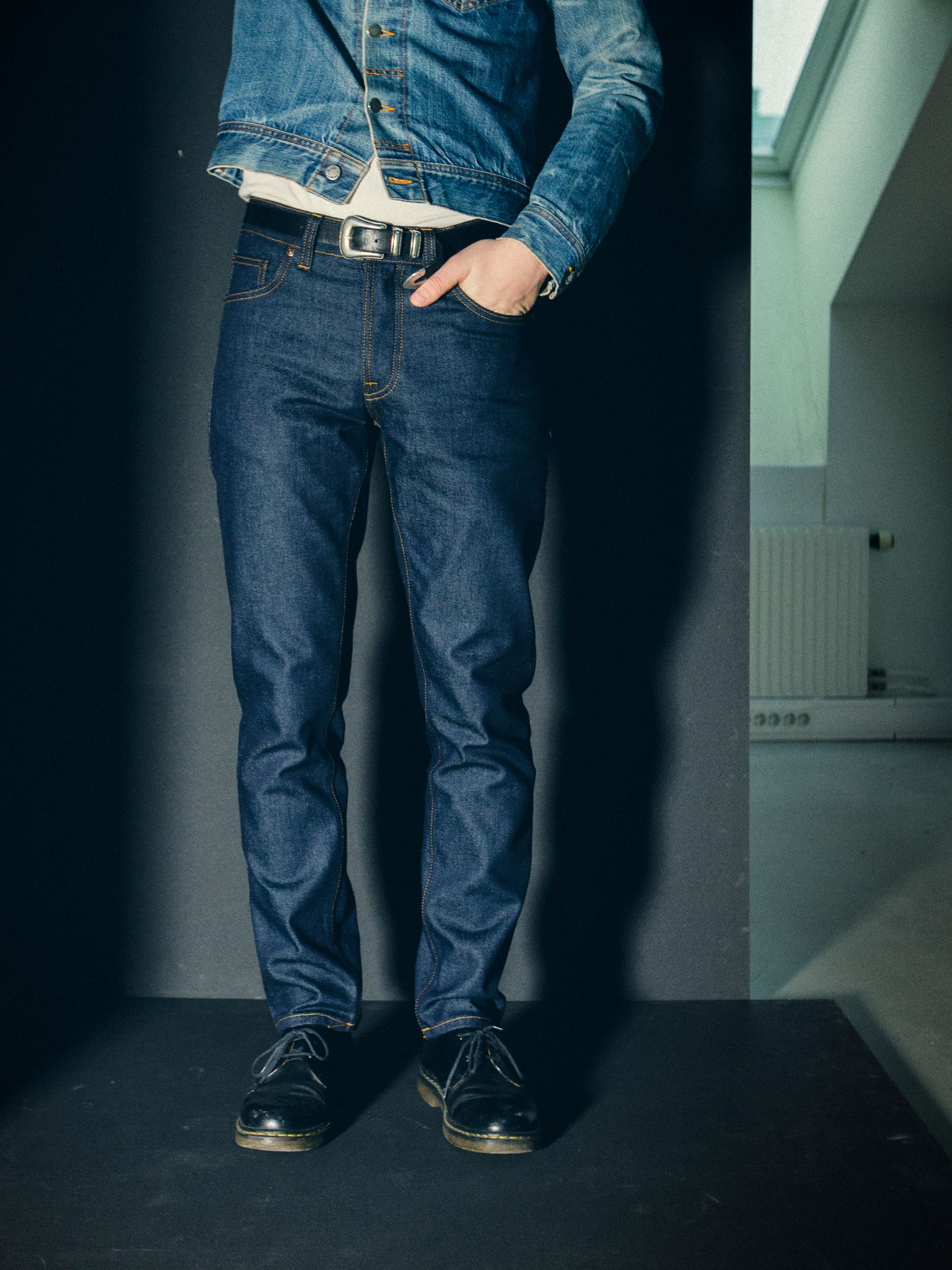 Nudie Jeans Solid Ollie Dry Abyss, men's slim fit jeans in dark blue, worn by a model against a dark wall.