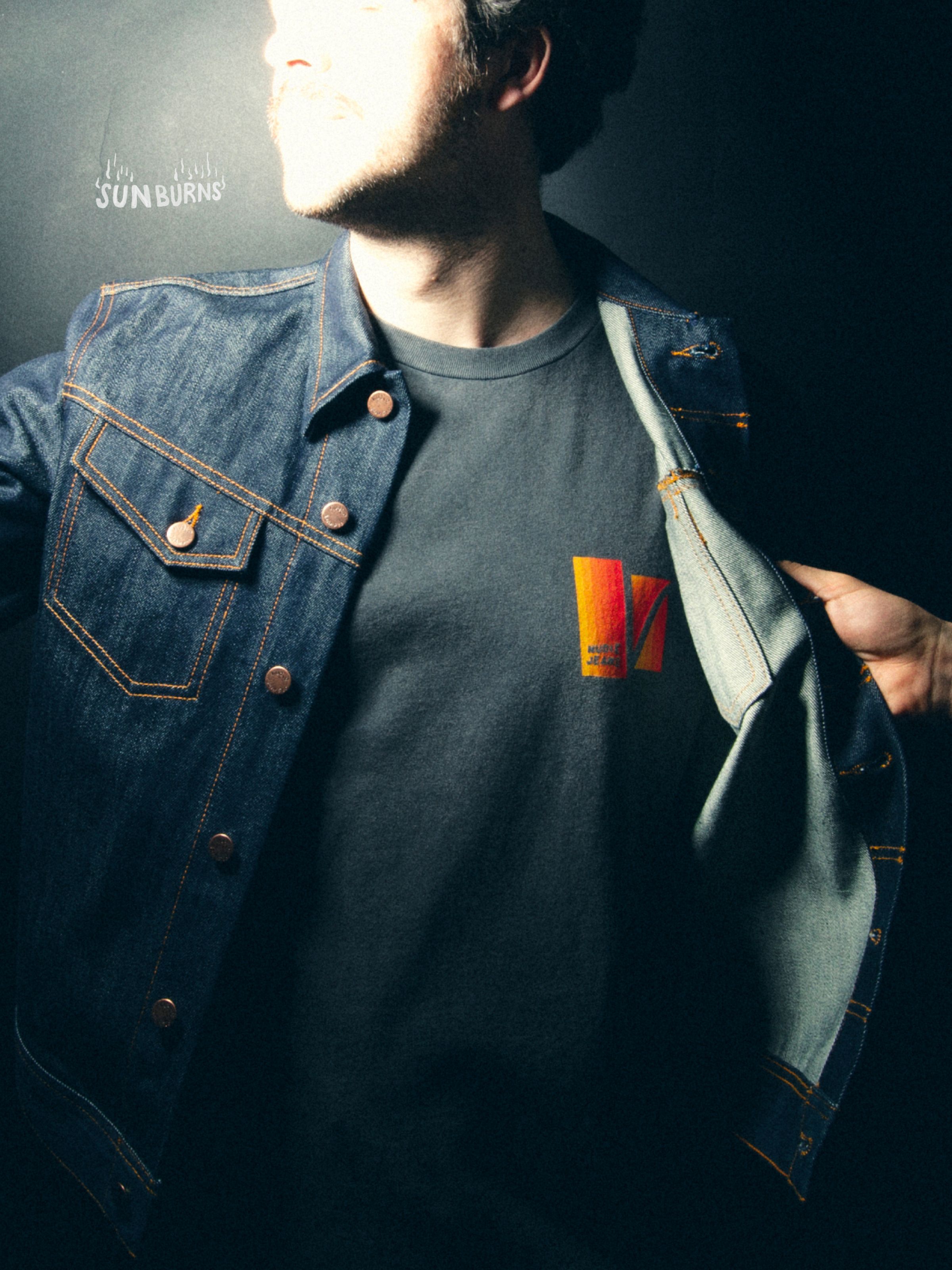 Nudie Jeans Roy Sunburns T-Shirt in Antracite, a men's tee worn by a model under a denim jacket, showing the chest print.