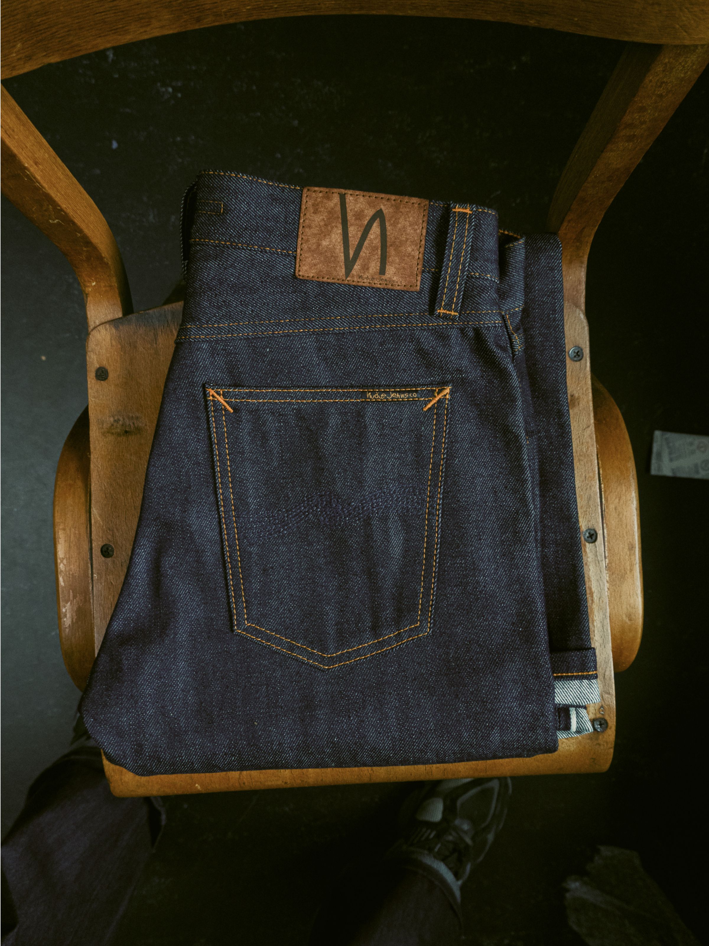 Nudie Jeans Steady Eddie II Dry Dusk Selv, reguläre Tapered-Herrenjeans aus Dry Selvage Denim, auf einem Holzstuhl gefaltet.