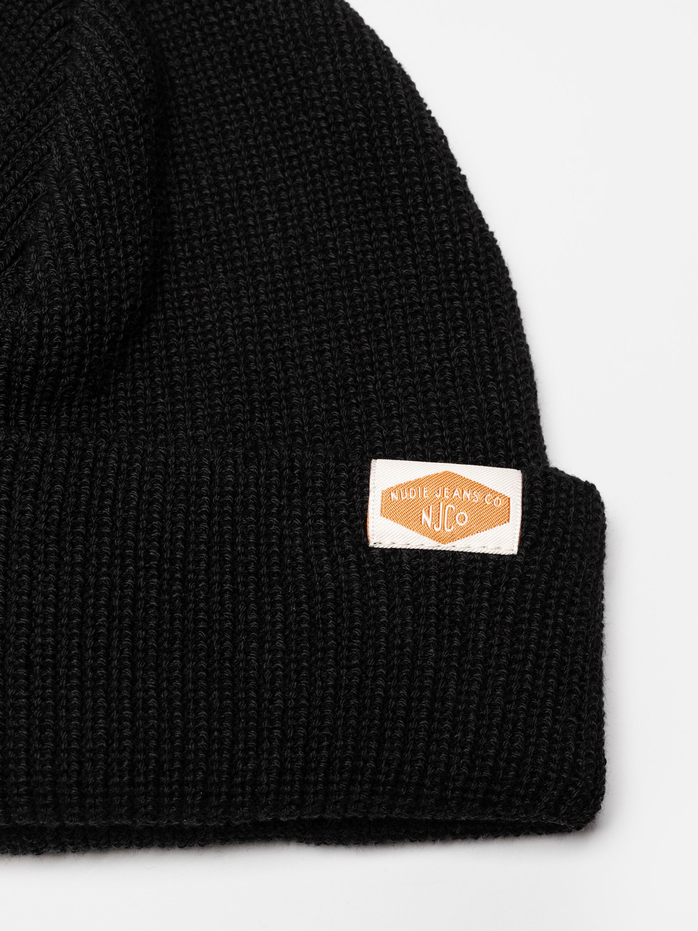 Nudie Jeans Cotton Beanie in black, a close-up showing the ribbed texture and the woven logo label on the folded cuff.