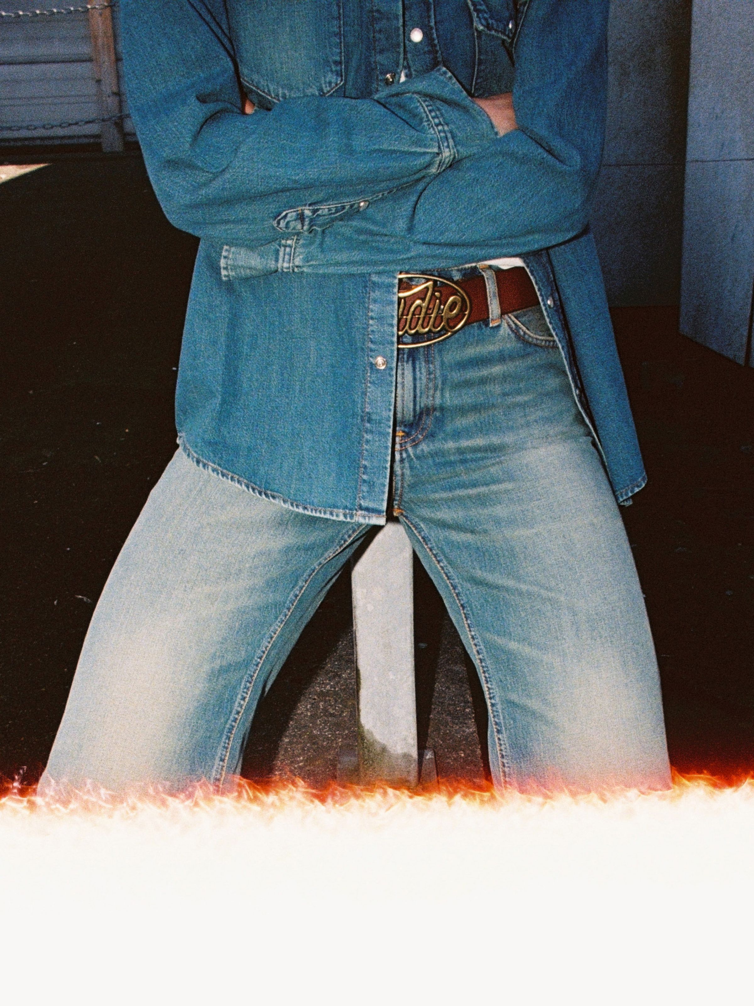 Nudie Jeans' Nudie Buckle Leather Belt in dark brown, worn by a model styled with a denim shirt and light-wash jeans.