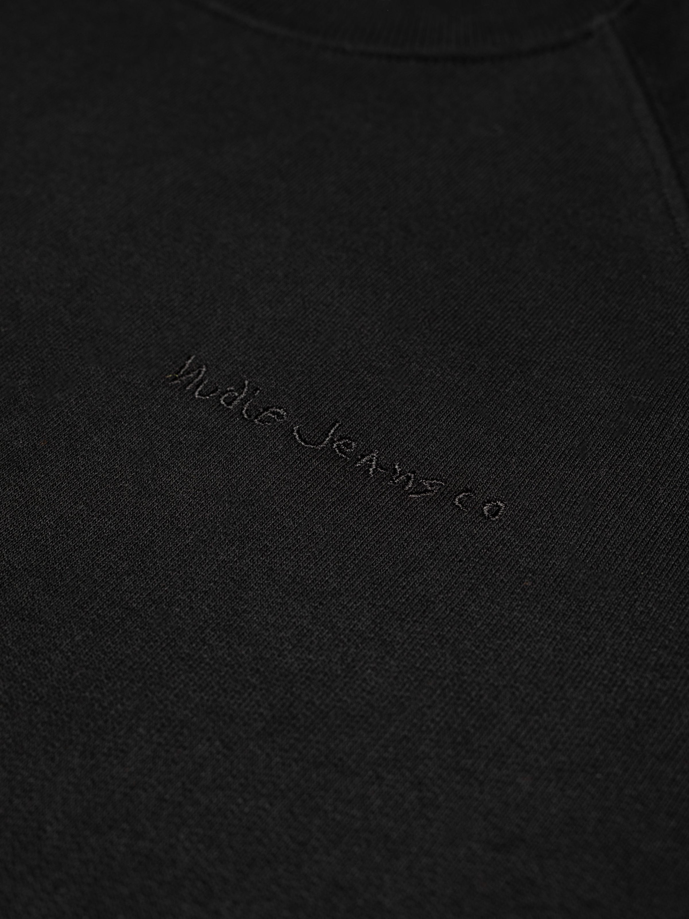 Nudie Jeans Palle Logo Sweatshirt in Black, a close-up view showing the tonal logo embroidery on the chest.