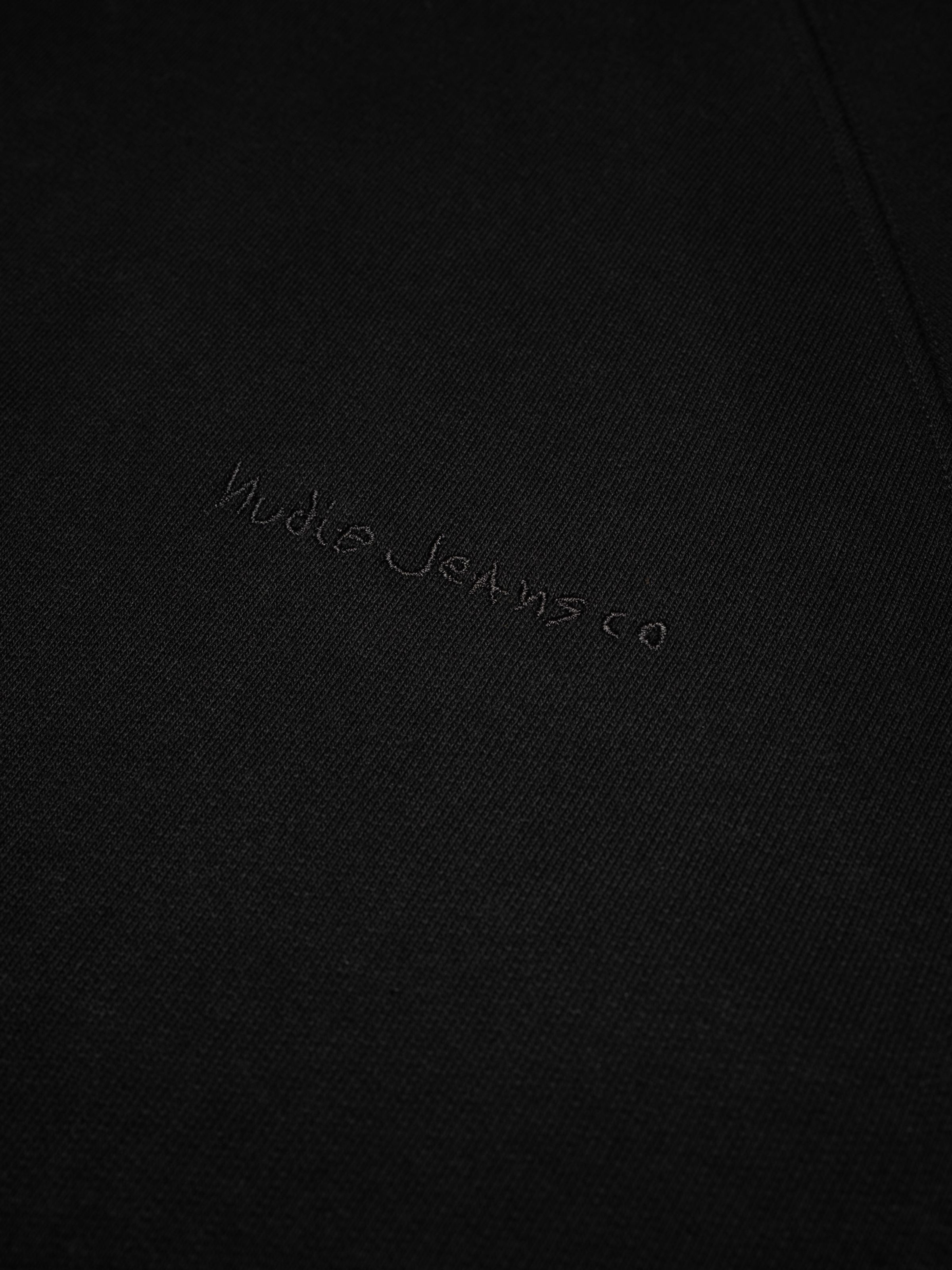 Nudie Jeans Gunde Logo Hoodie in Black, a close-up detail of the tone-on-tone logo embroidery on the chest.