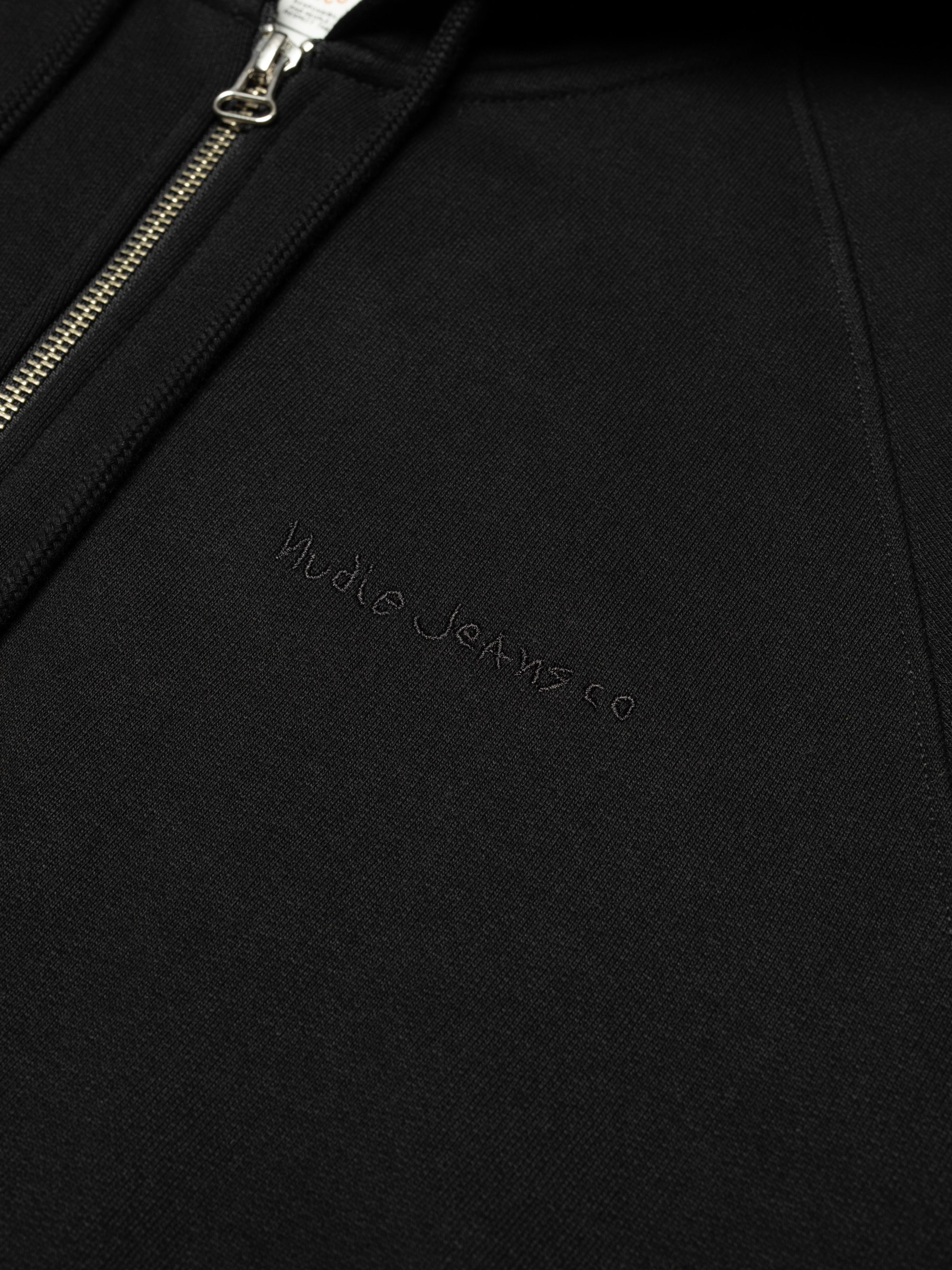 Nudie Jeans Gunde Logo Zip Hoodie in Black, showing a close-up of the embroidered chest logo and zipper.