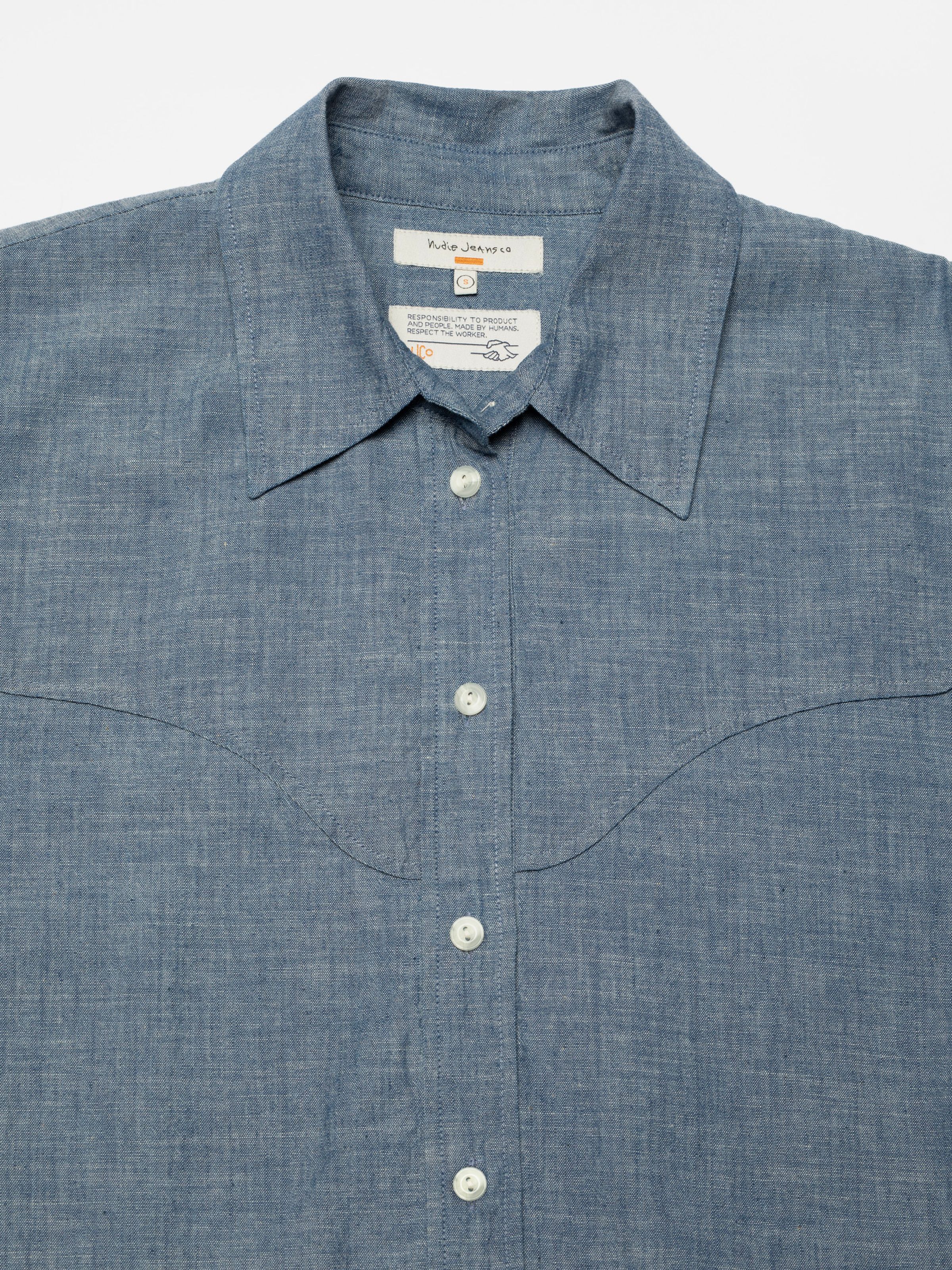 Nudie Jeans Daisy Chambray Western Shirt, a women's shirt in blue, shown laid flat in a close-up of the collar and yoke.