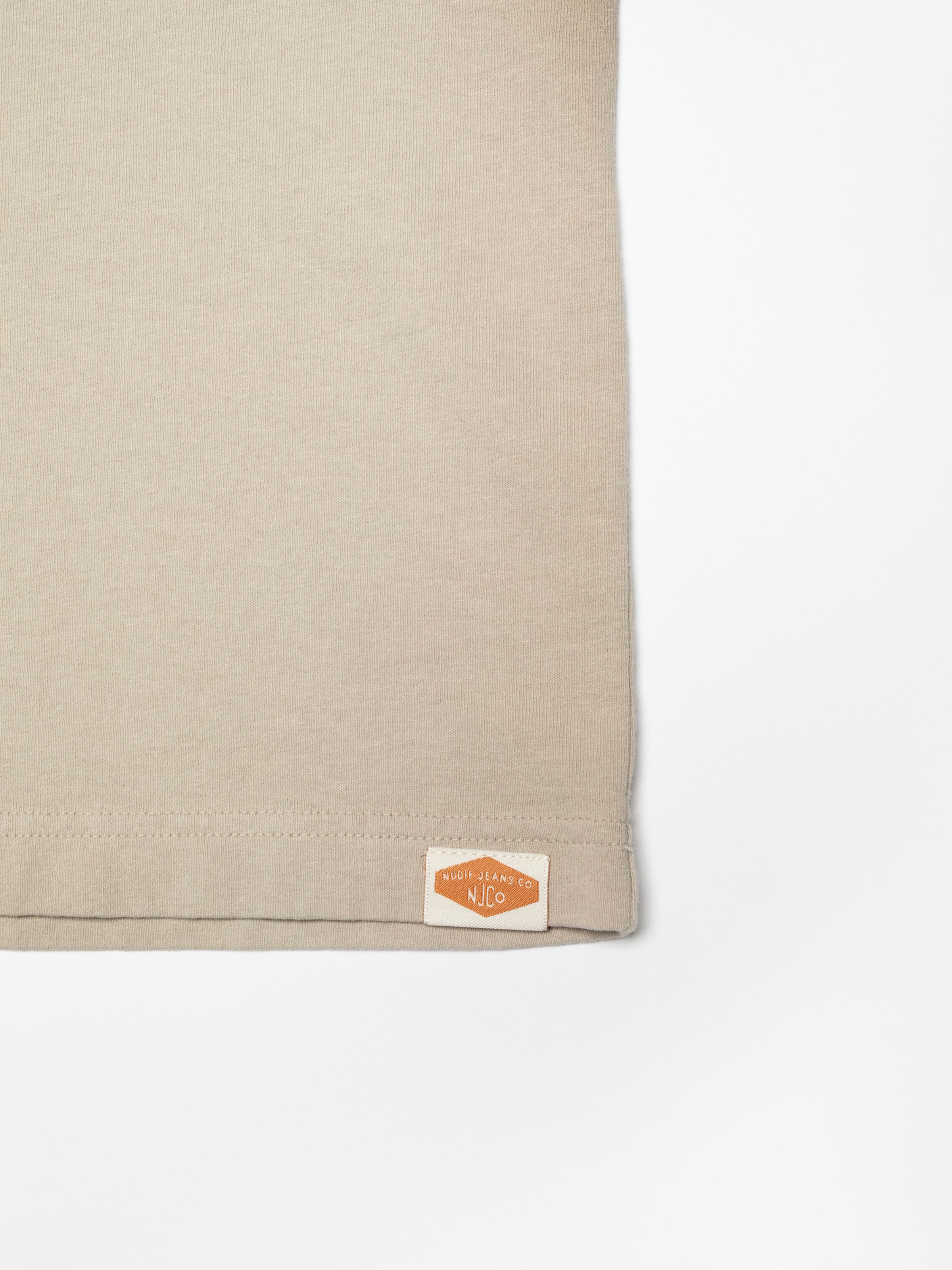 Nudie Jeans Uno Everyday T-Shirt in Sand, close-up of the bottom hem showing the woven logo label.