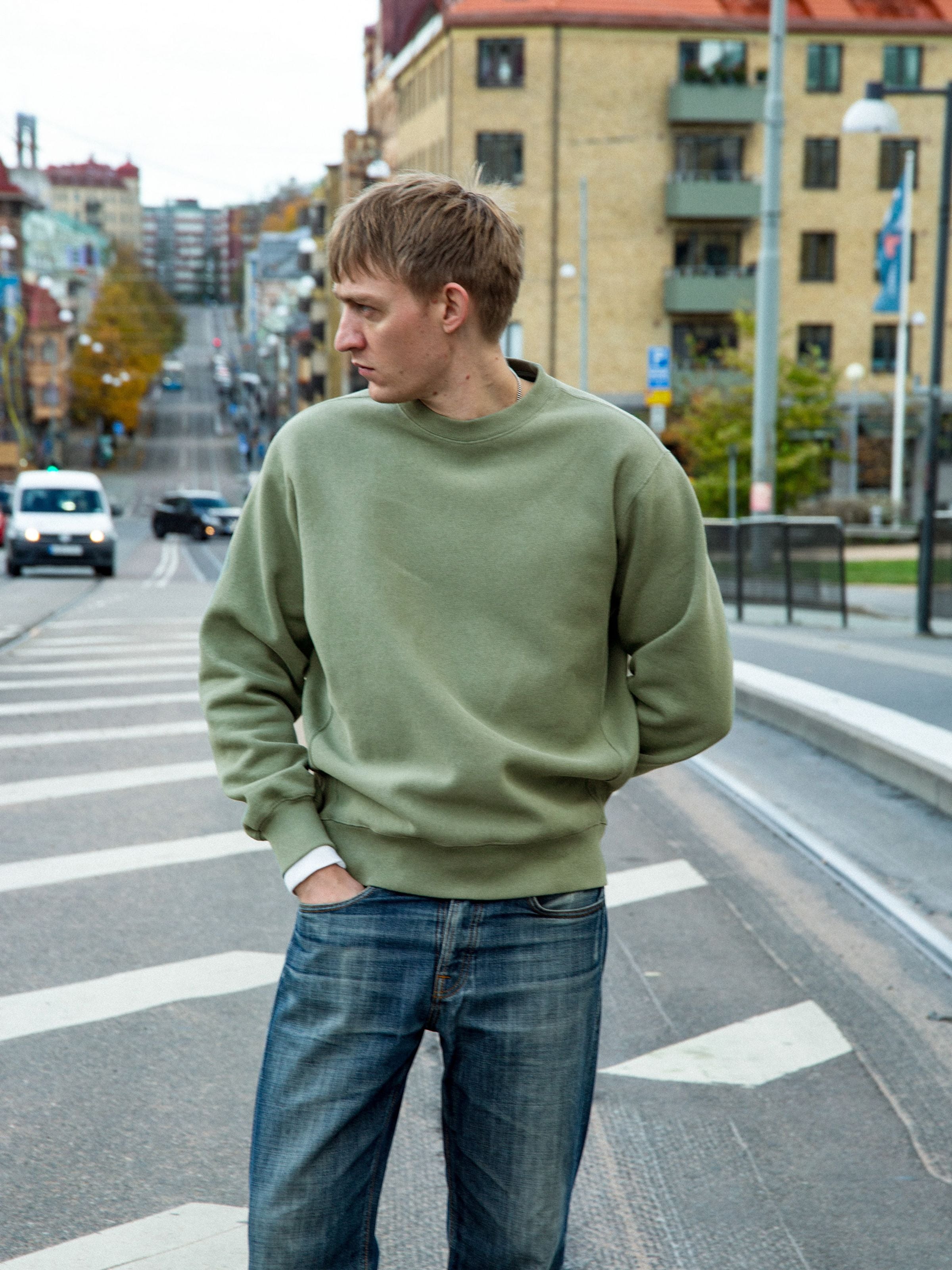 Nudie Jeans Hasse Crew Neck Sweatshirt in green, shown on a male model standing on a city street.