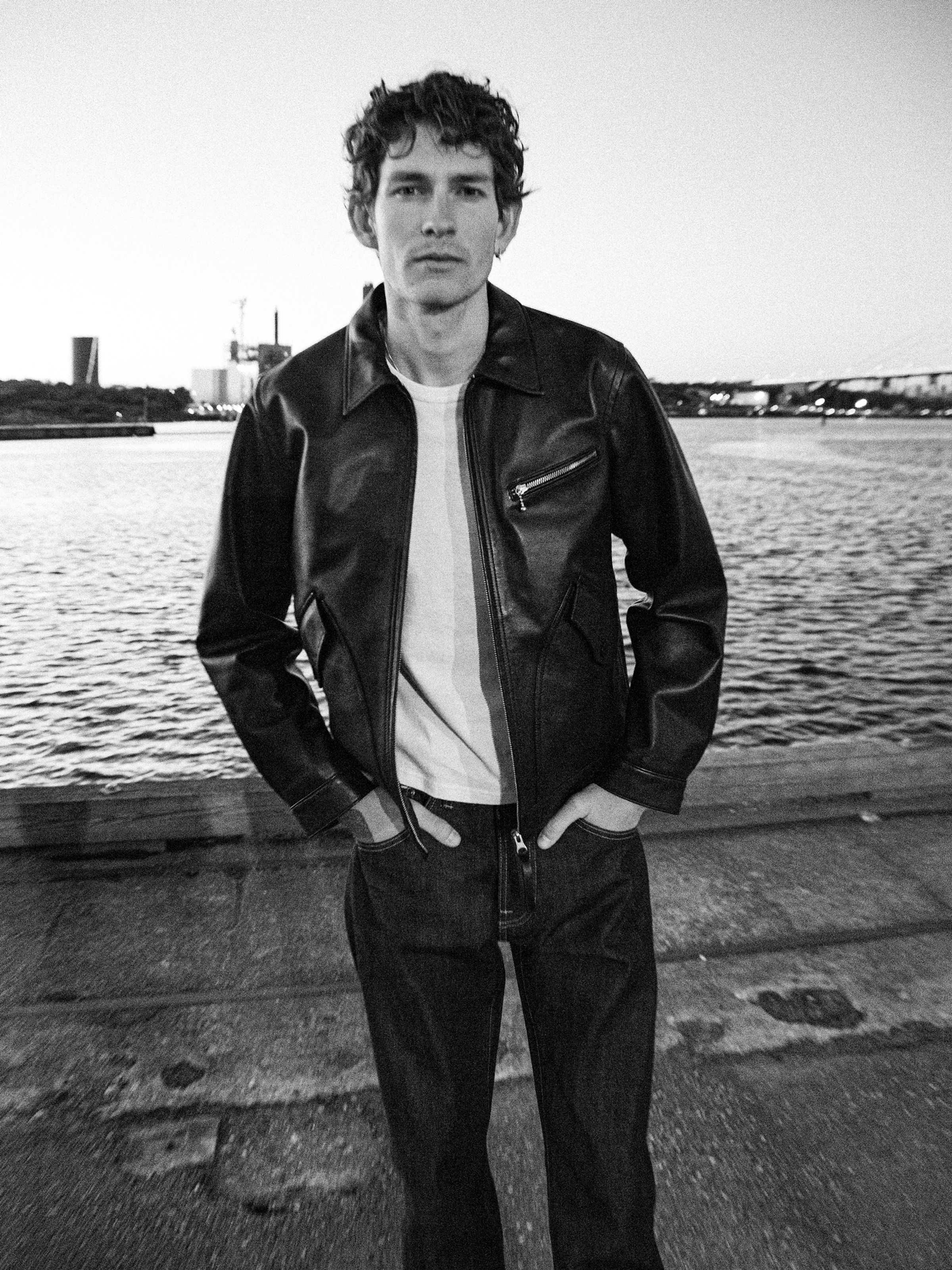 Nudie Jeans Blake Zip Jacket Leather in Black, a men's regular fit jacket, shown on a model in an outdoor waterside setting.