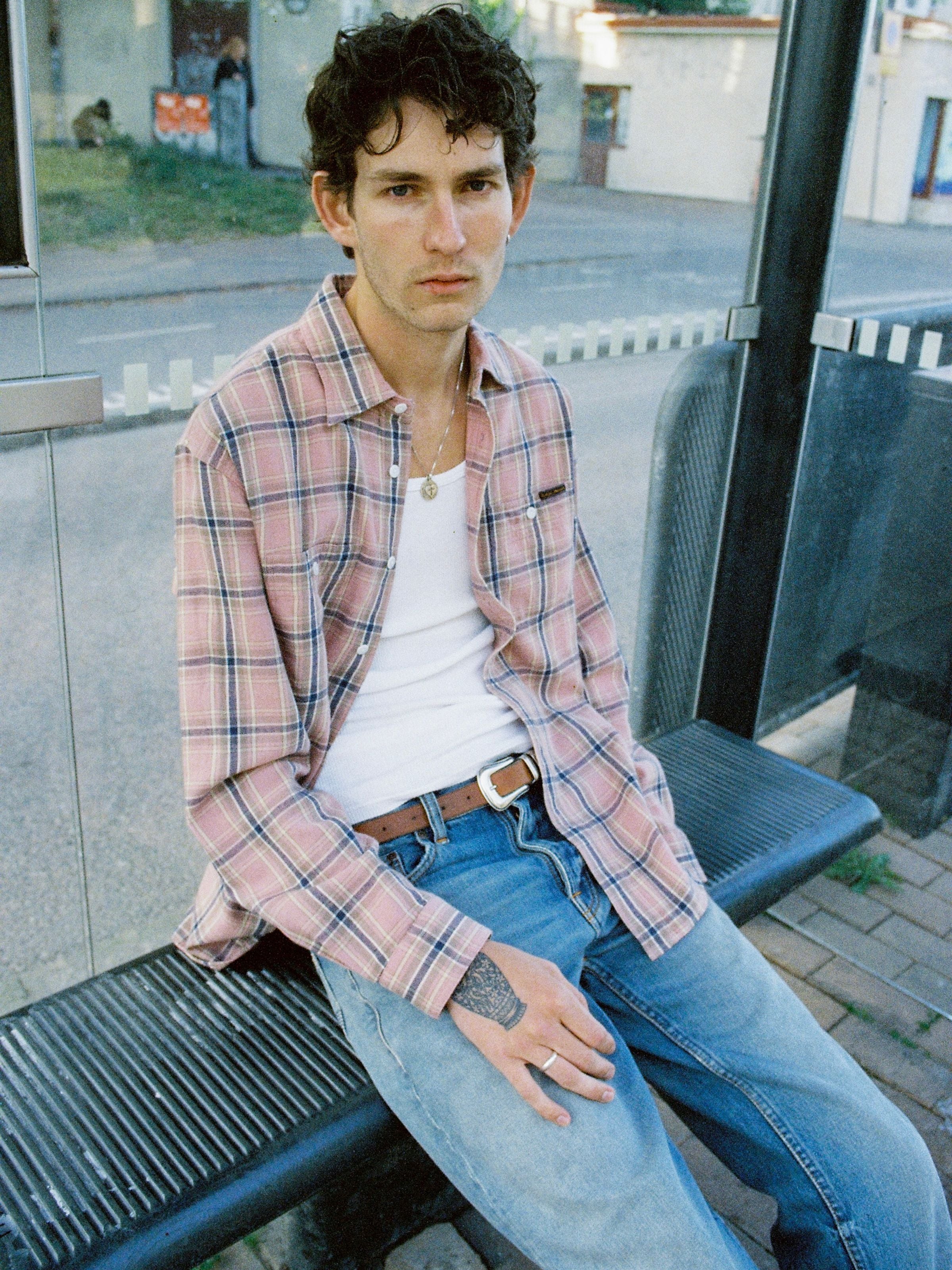 Nudie Jeans Julian Worker Shirt Checked in Pink, men's regular fit shirt shown on a model sitting at a bus stop.