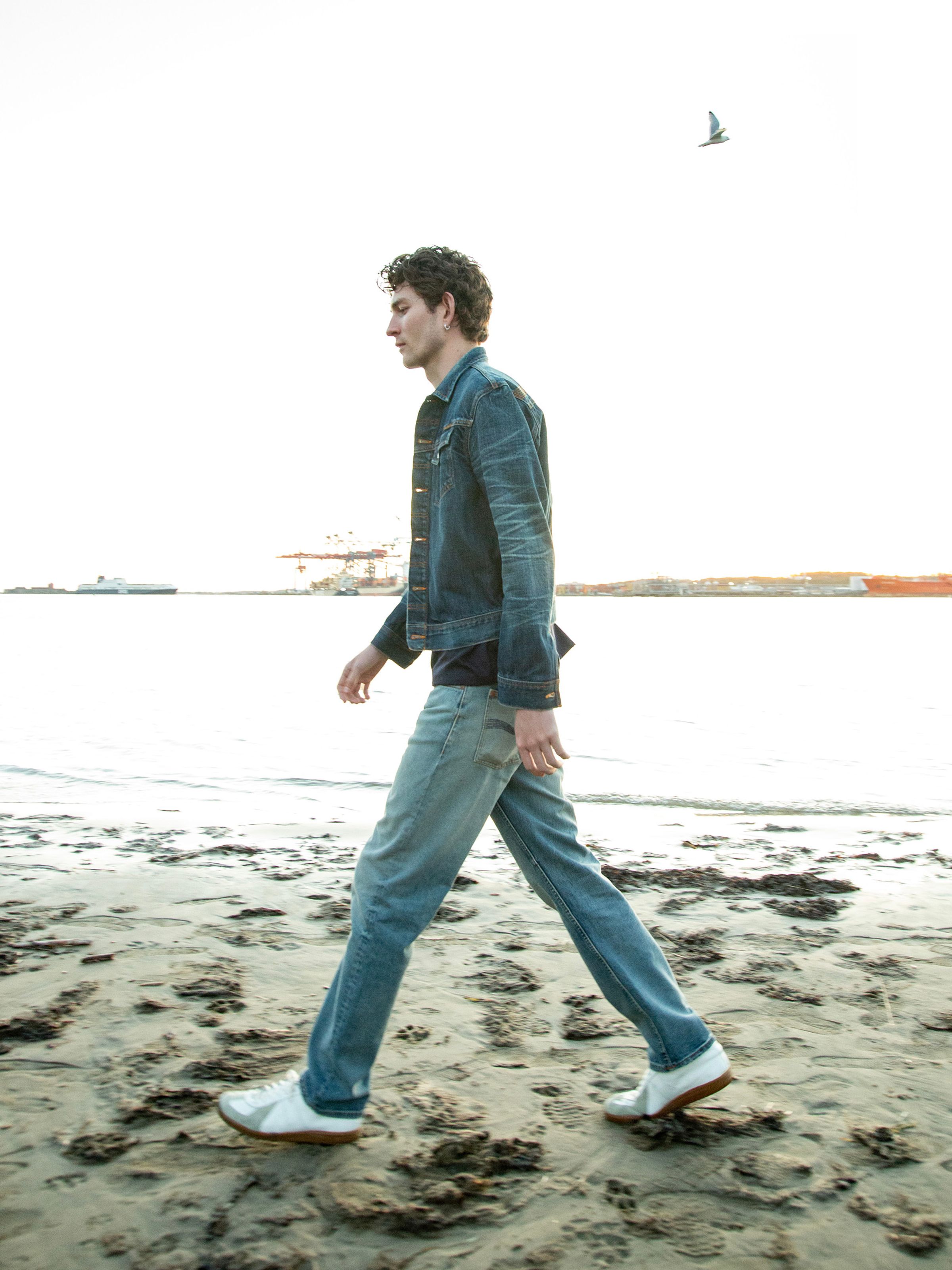 Nudie Jeans Conny Denim Jacket Heavy Used, a slim fit denim jacket shown from the side on a model walking on a beach.