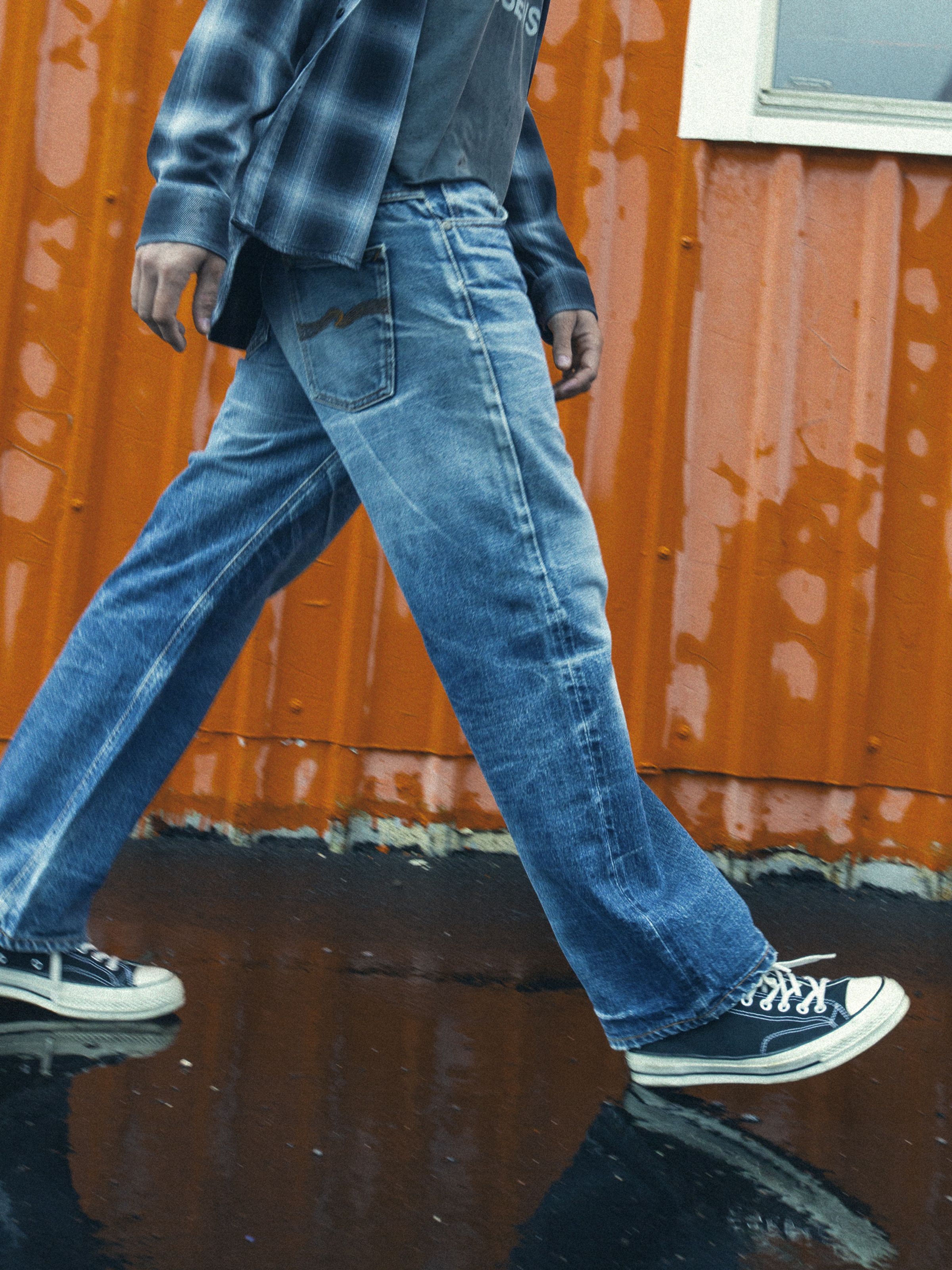 Nudie Jeans Tuff Tony Mystery Original Life, a men's loose fit jean, worn by a model walking past an orange wall.