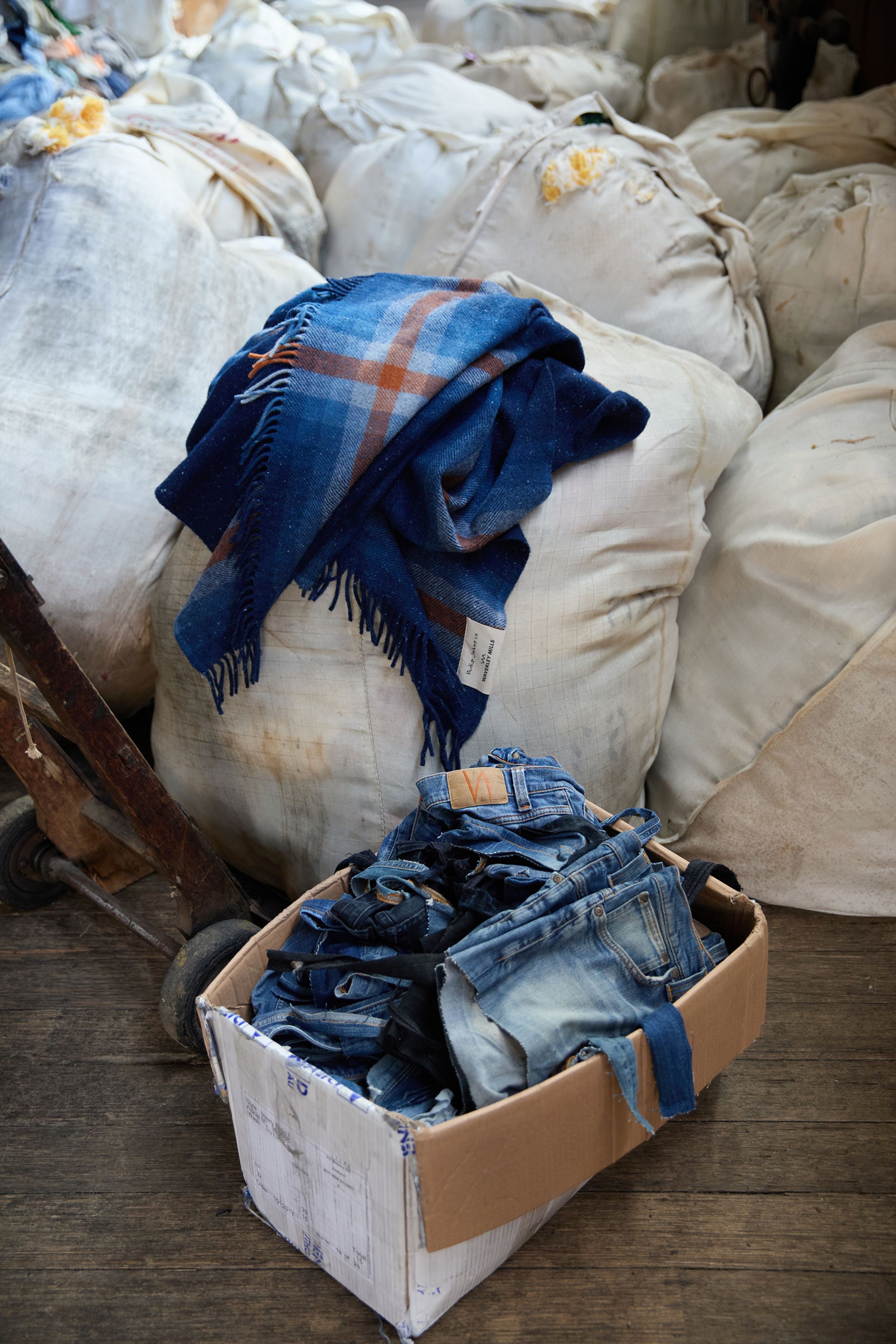 Nudie Jeans NJ x WM Recycled Blanket, a blue plaid blanket, shown in a workshop alongside a box of recycled denim jeans.