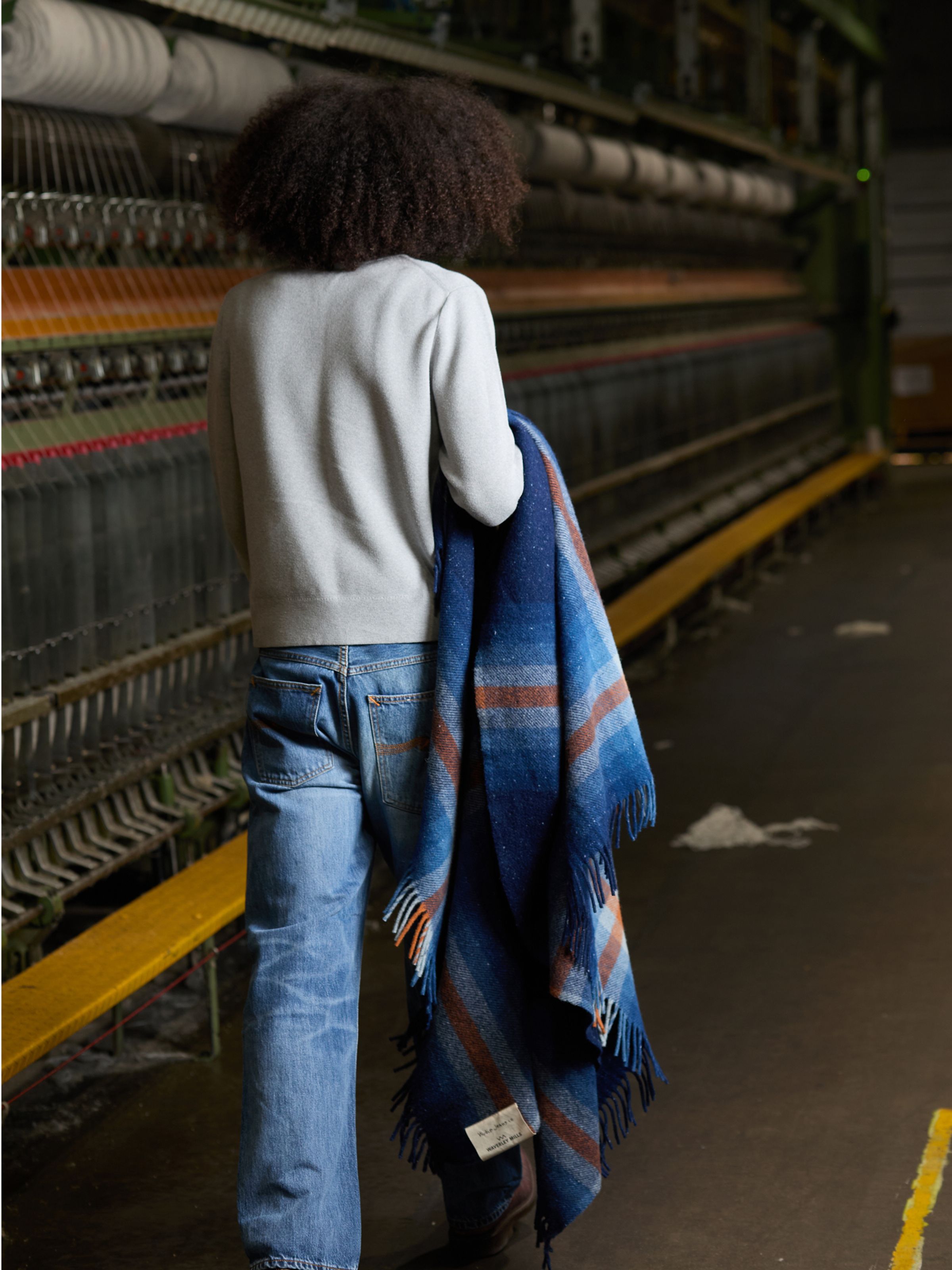 Nudie Jeans NJ x WM Recycled Blanket, a blue recycled wool blanket, held by a model shown from the back in a textile mill.