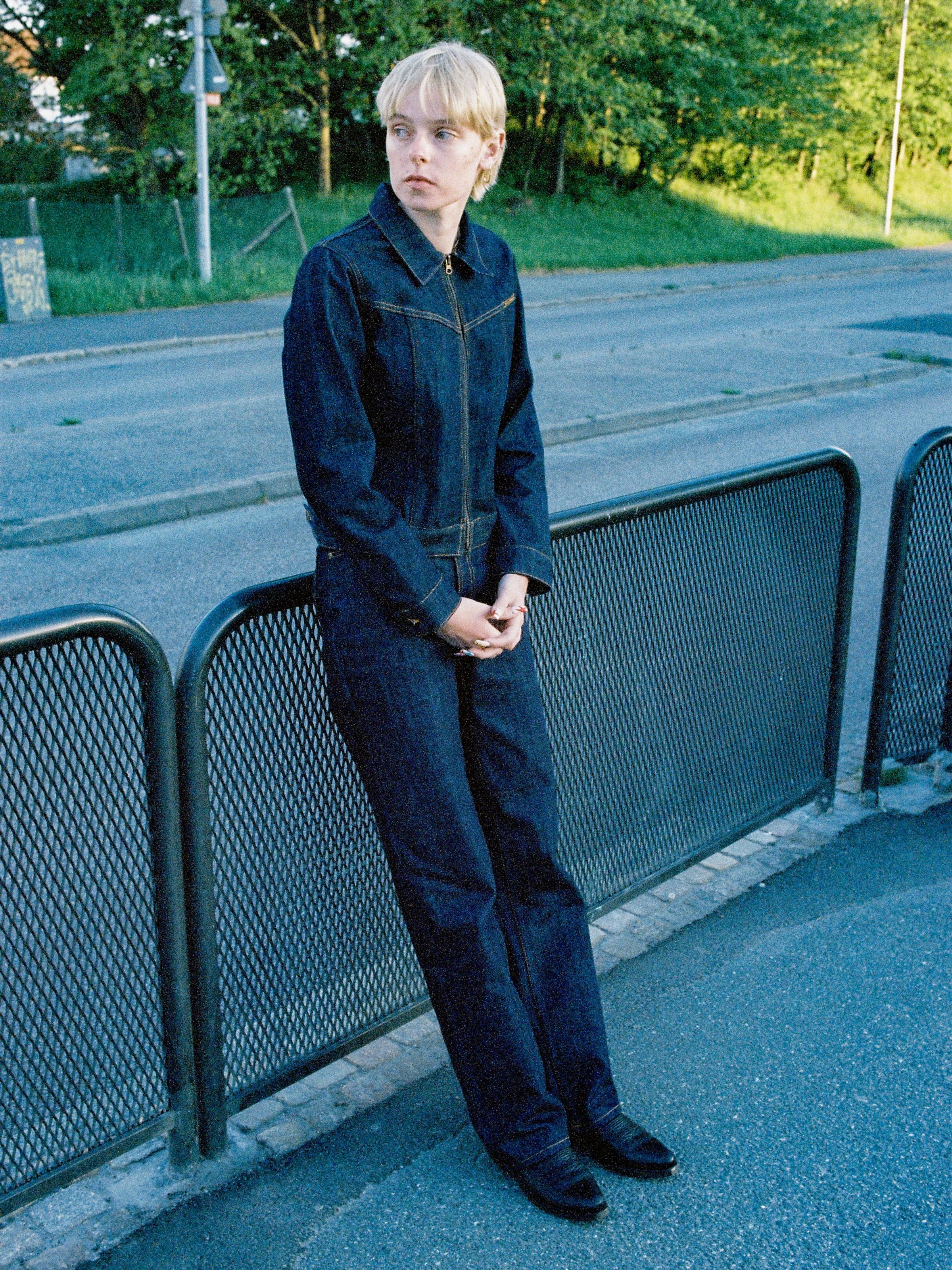 Nudie Jeans Britta Denim Jacket Dry Streaky, a women's slim-fit indigo denim jacket on a model leaning against a metal fence.