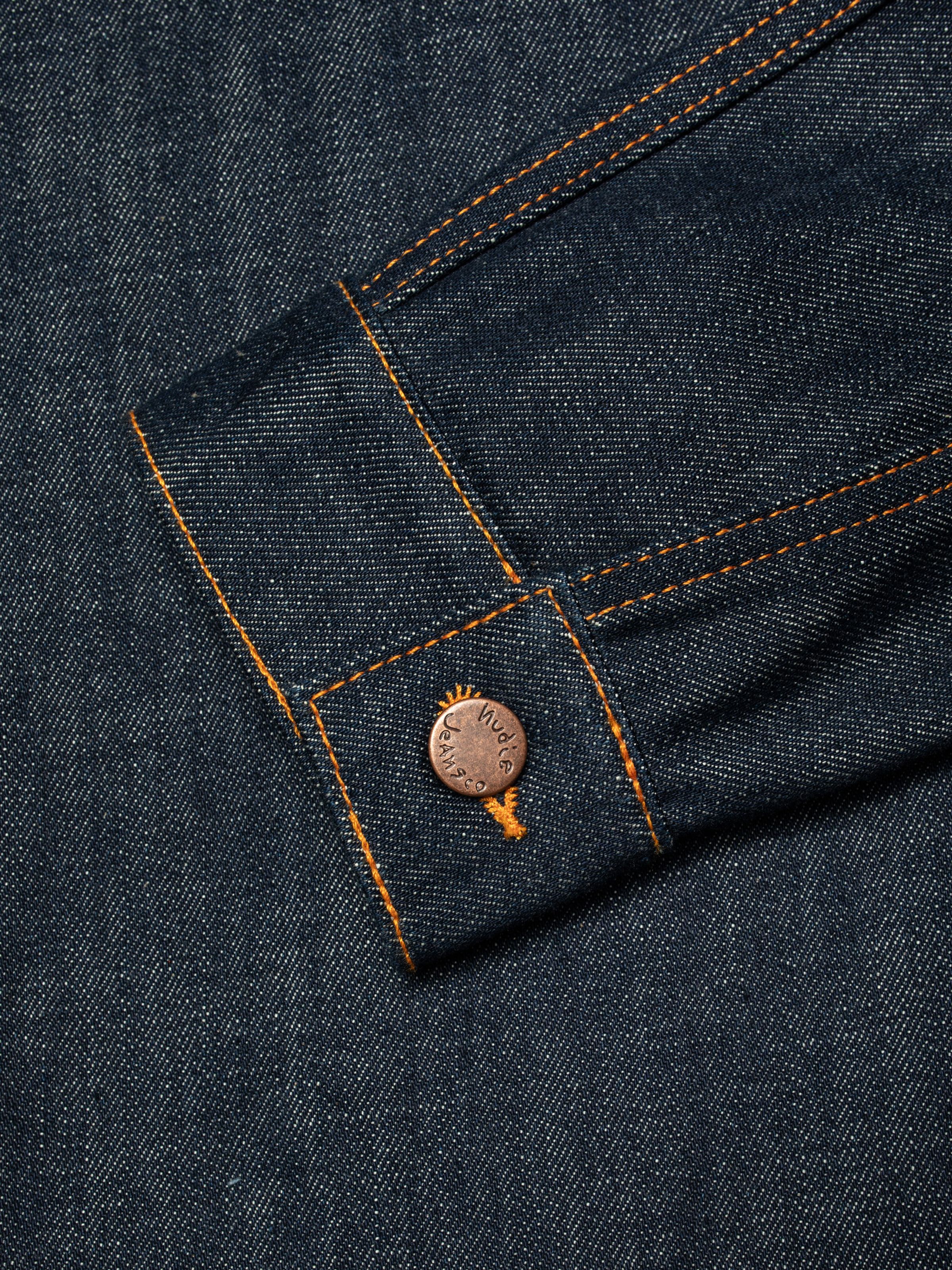 Nudie Jeans Britta Denim Jacket Dry Streaky, a close-up of the sleeve cuff with branded copper button and orange stitching.