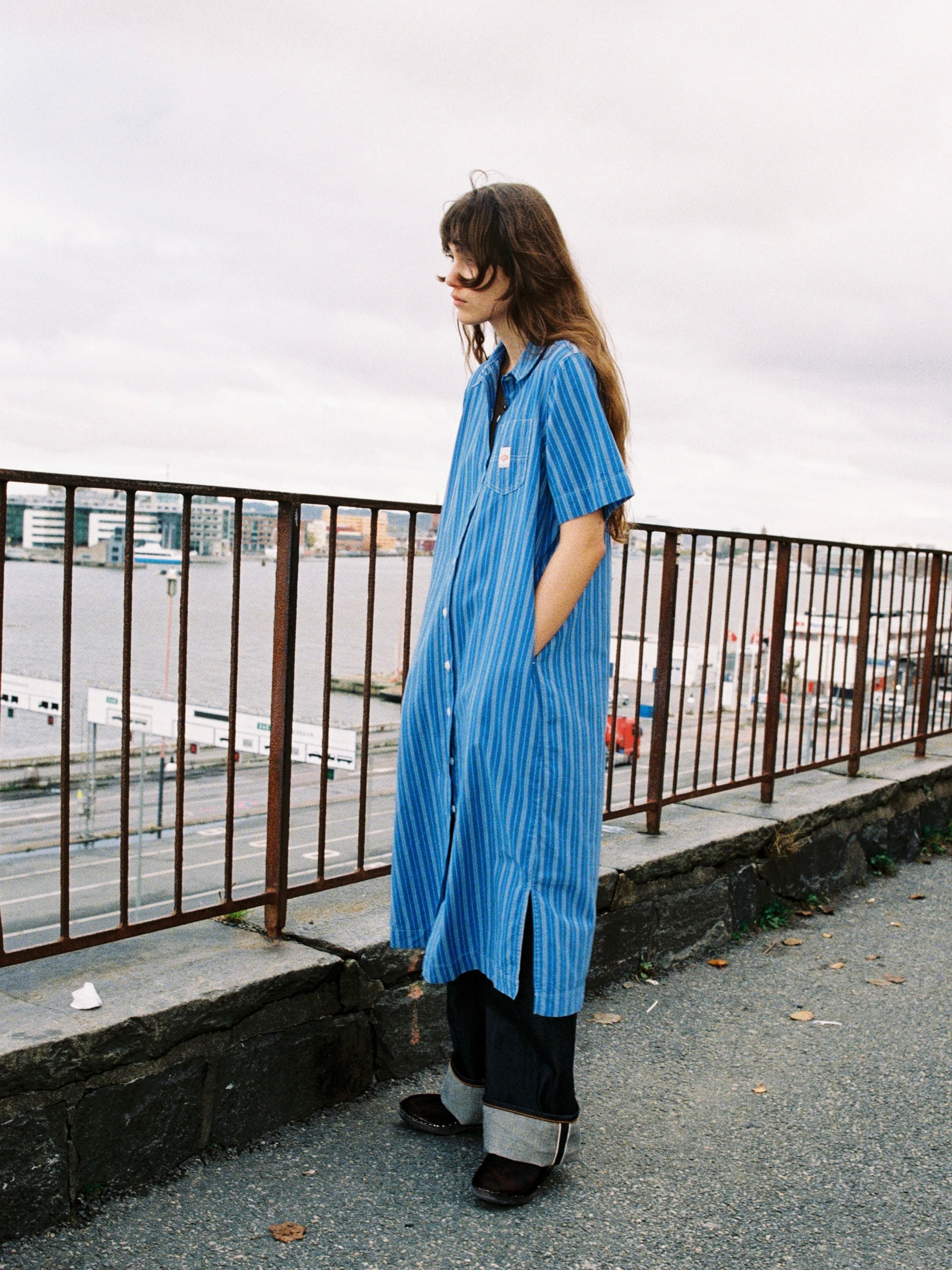Nudie Jeans Danielle Dress Striped, ein blau-cremeweiß gestreiftes Damen-Hemdblusenkleid, an einem Model am Wasser gezeigt.