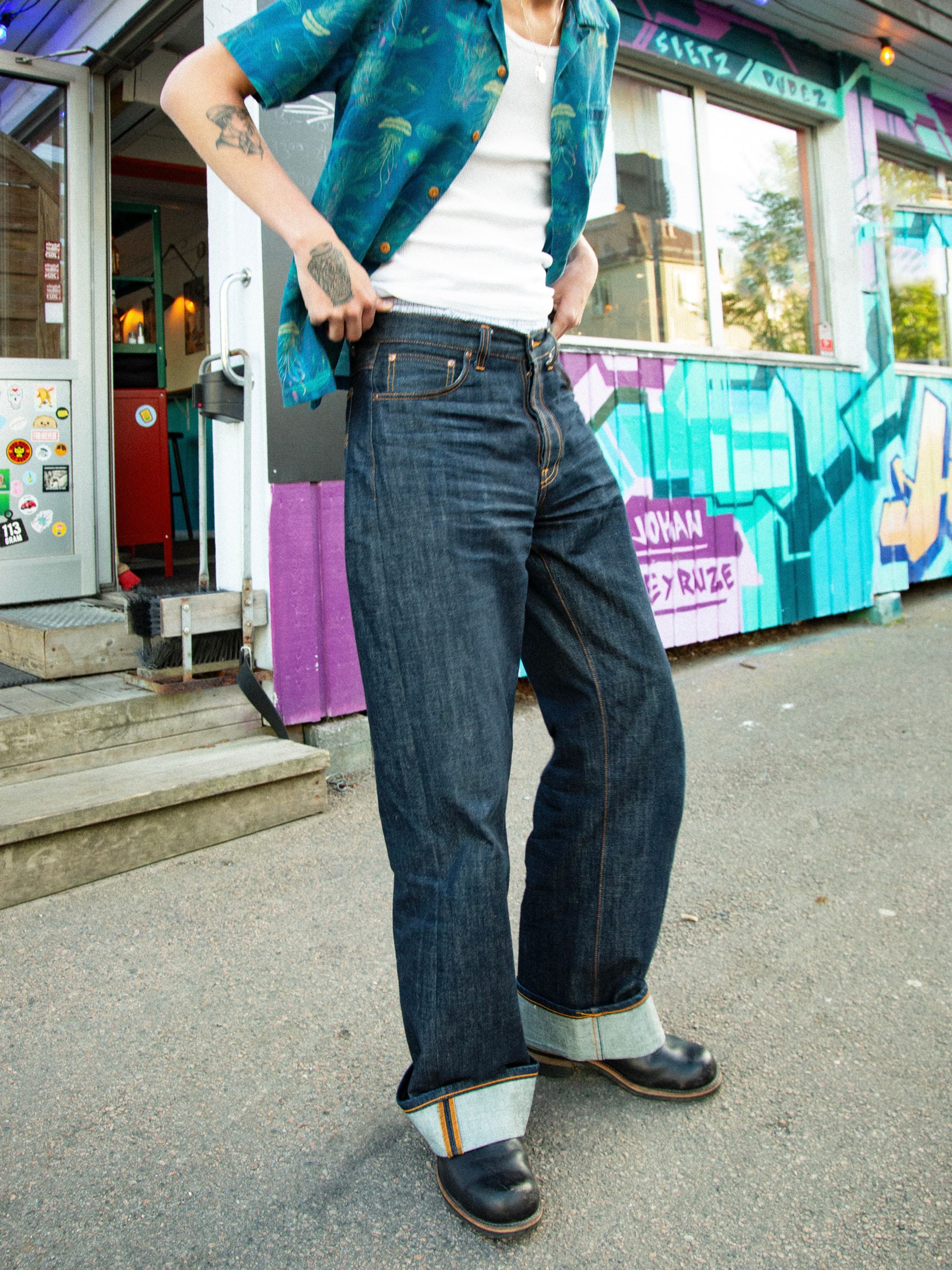 Nudie Jeans Loud Larry Dry Streaky, lockere Herrenjeans an einem Model im Freien mit breiten, umgeschlagenen Selvedge-Säumen.