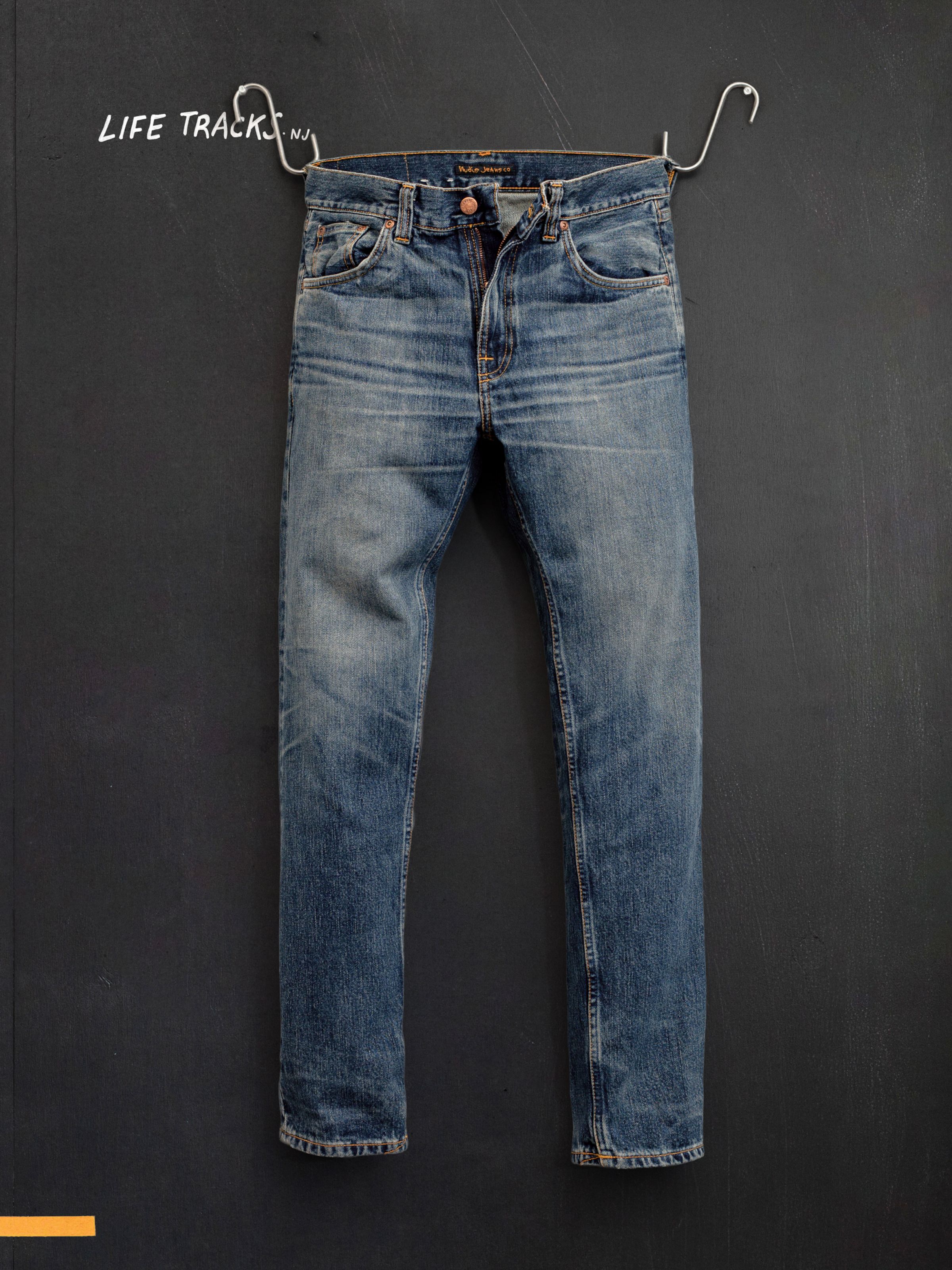 Nudie Jeans Gritty Jackson Sand Tracks, men's regular fit jeans, shown in a flatshot hanging against a dark wall.