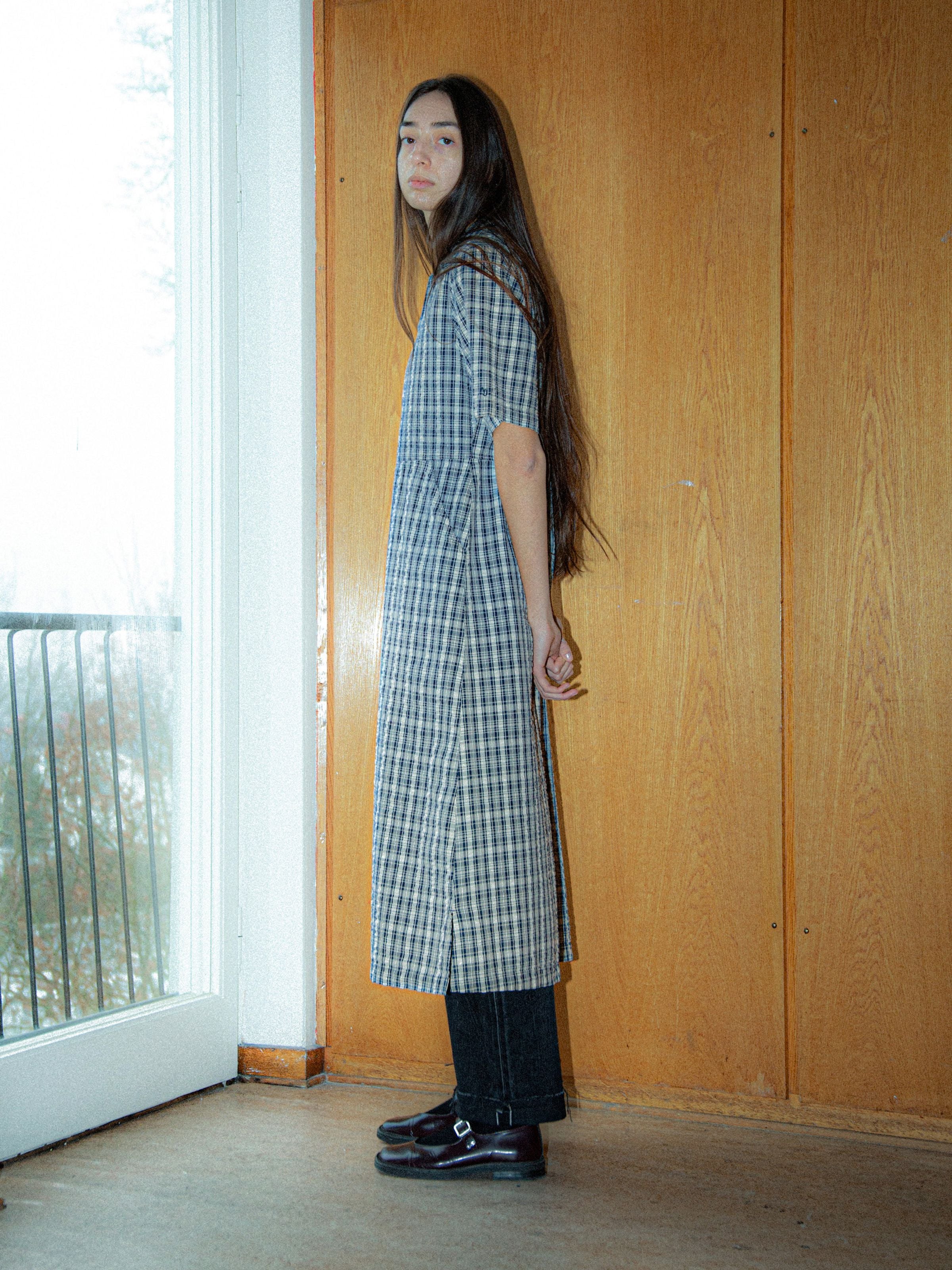 Nudie Jeans Ronja Dress Indigo Seersucker, kurzärmeliges Damenkleid, von der Seite an einem Model vor einer Holzwand.