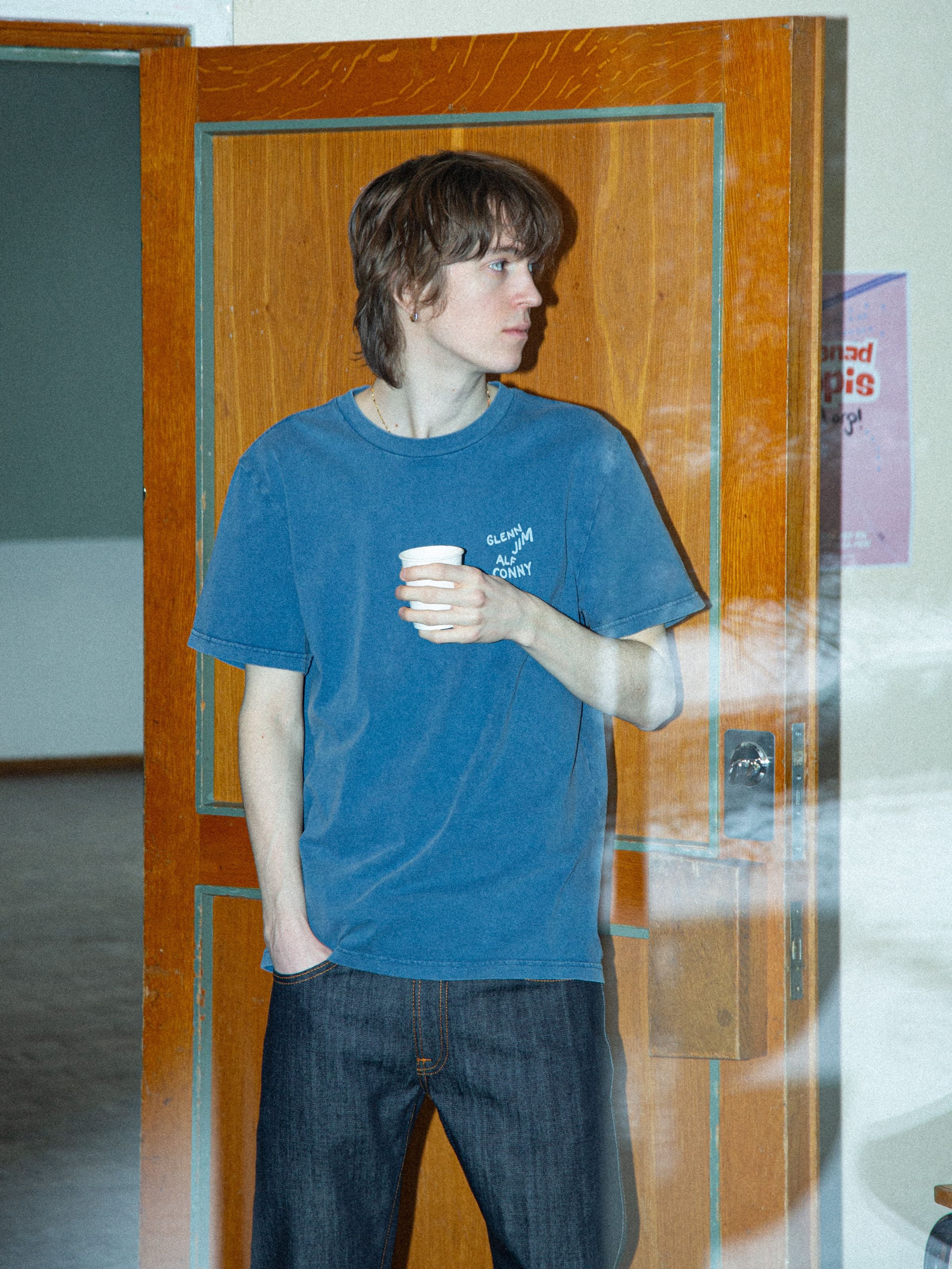 Nudie Jeans Roy T-Shirt OG Line-Up French Blue, lifestyle shot of a model wearing the tee in front of a wooden door.