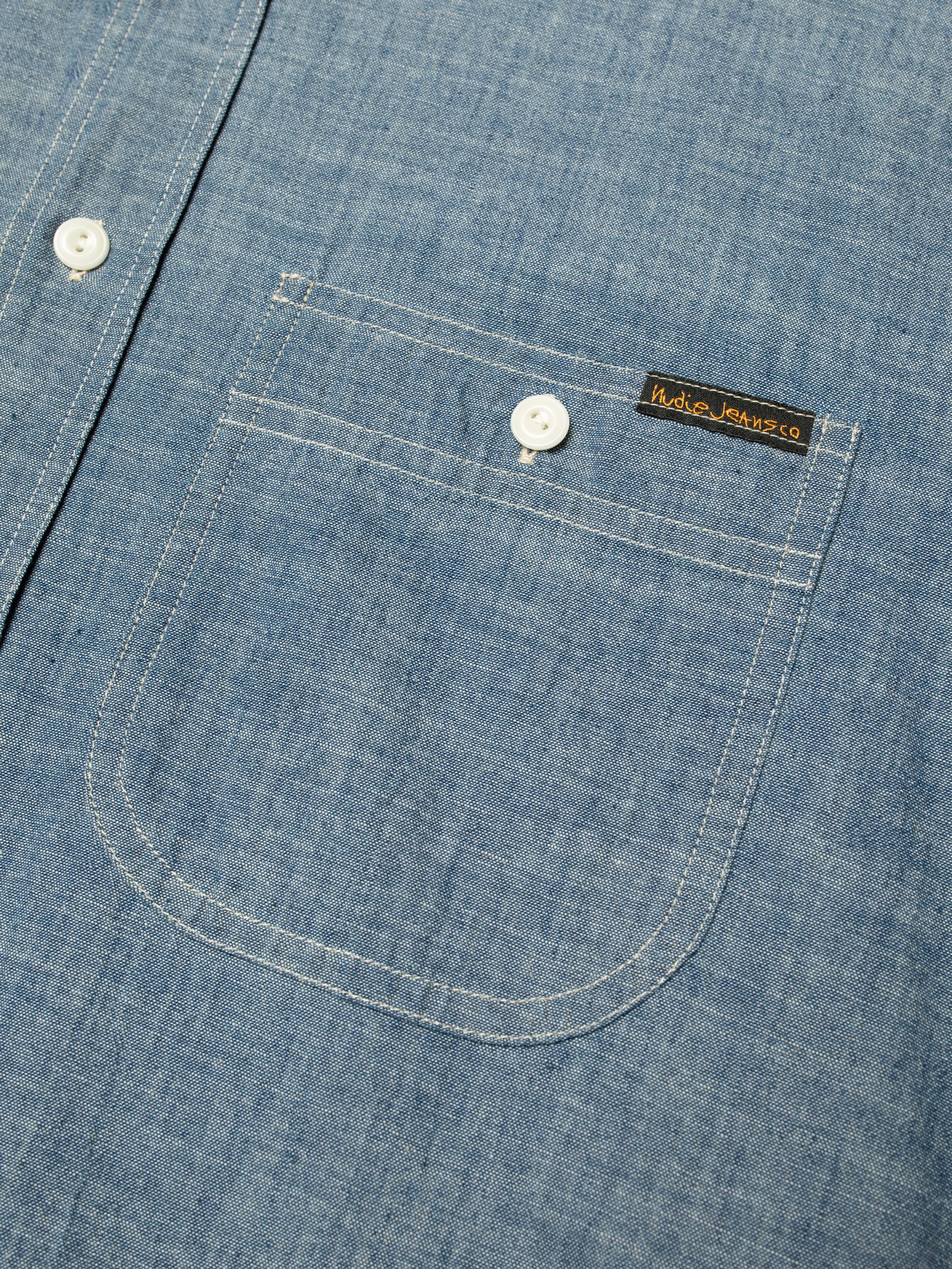 Nudie Jeans Julian Worker Shirt Chambray, close-up of the chest pocket showing the white stitching and logo label.