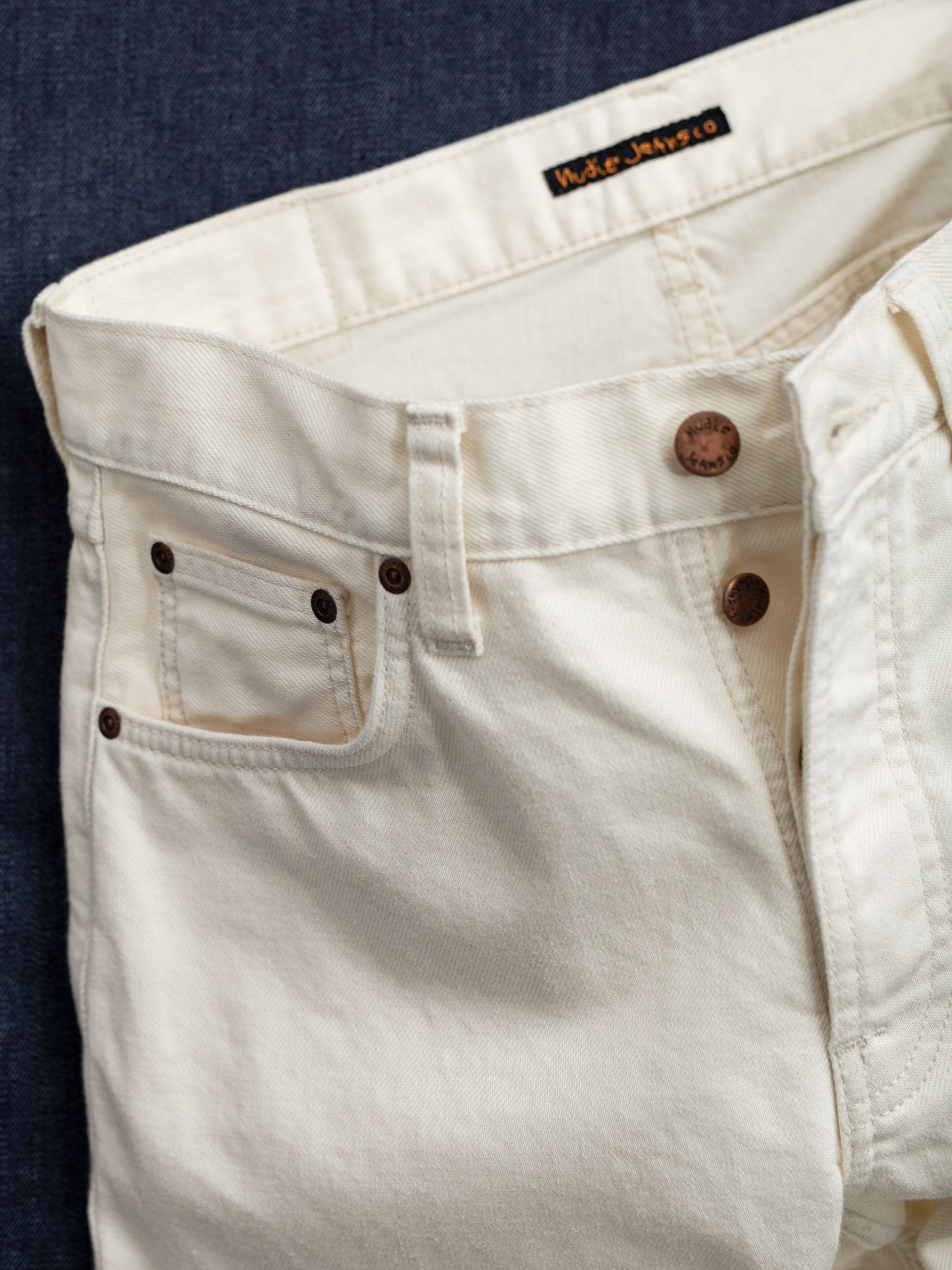 Nudie Jeans Solid Ollie Everivory, Nahaufnahme des Comfort Stretch Denim in Ivory mit Kupferknöpfen und Button Fly.