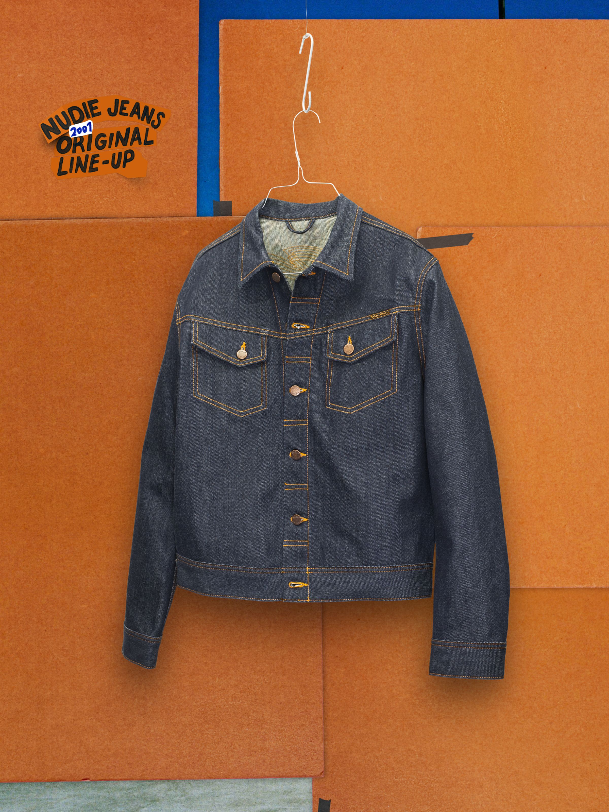 Nudie Jeans Conny Denim Jacket Dry One, a slim-fit dark blue rigid denim jacket with orange stitching on a brown backdrop.