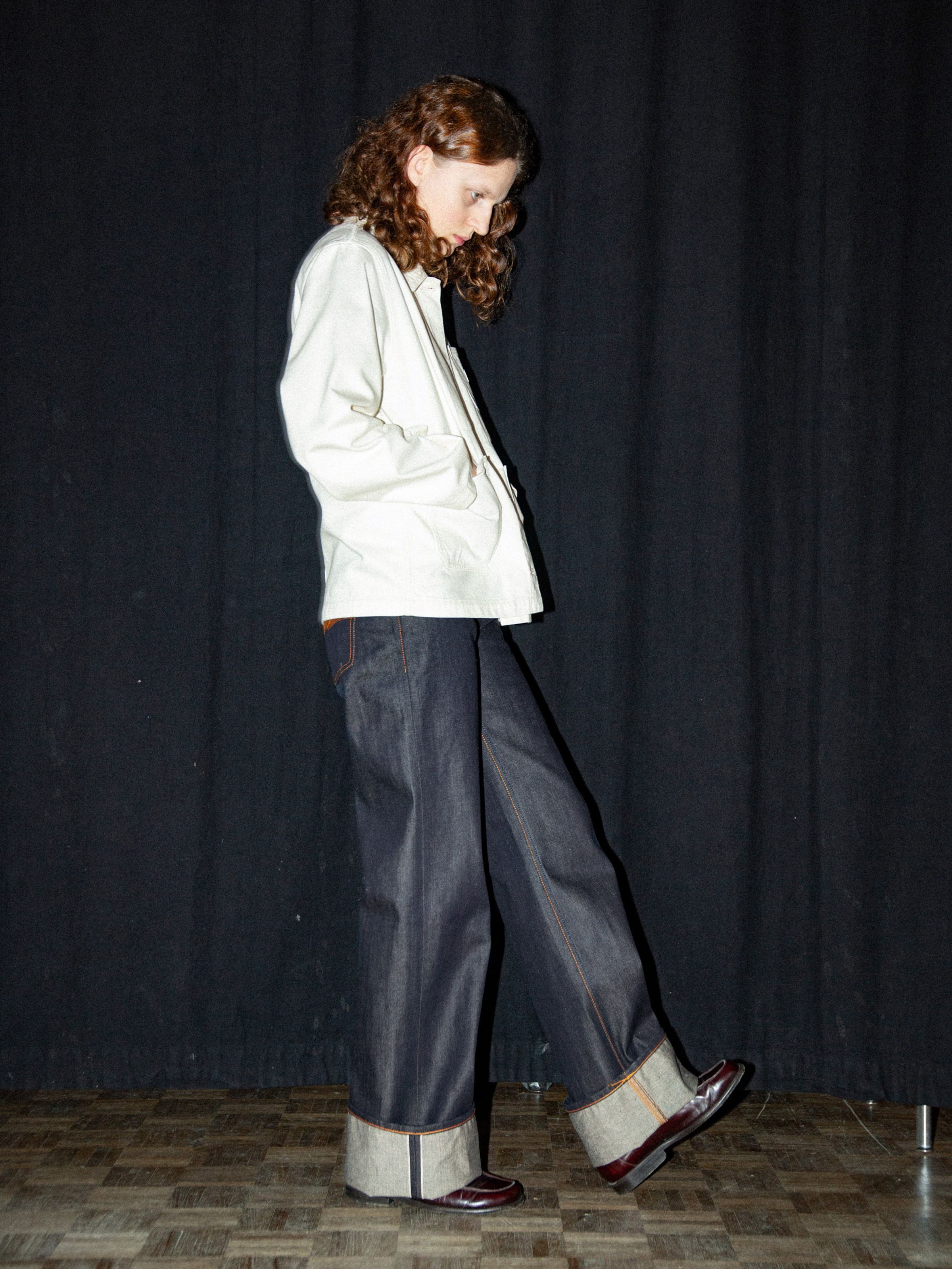 Nudie Jeans Ada Workwear Jacket Ivory, Damenjacke am Model mit umgeschlagenen Selvedge-Jeans vor einem schwarzen Vorhang.