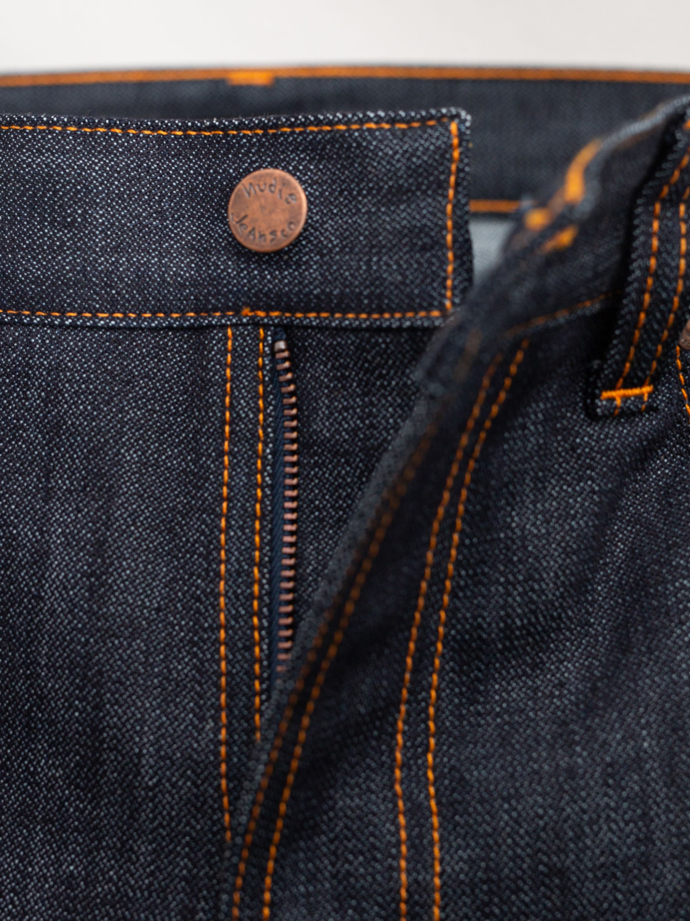 Nudie Jeans Slim Jim Dry Two, a close-up of the zip fly and copper logo button on indigo organic cotton broken twill denim.