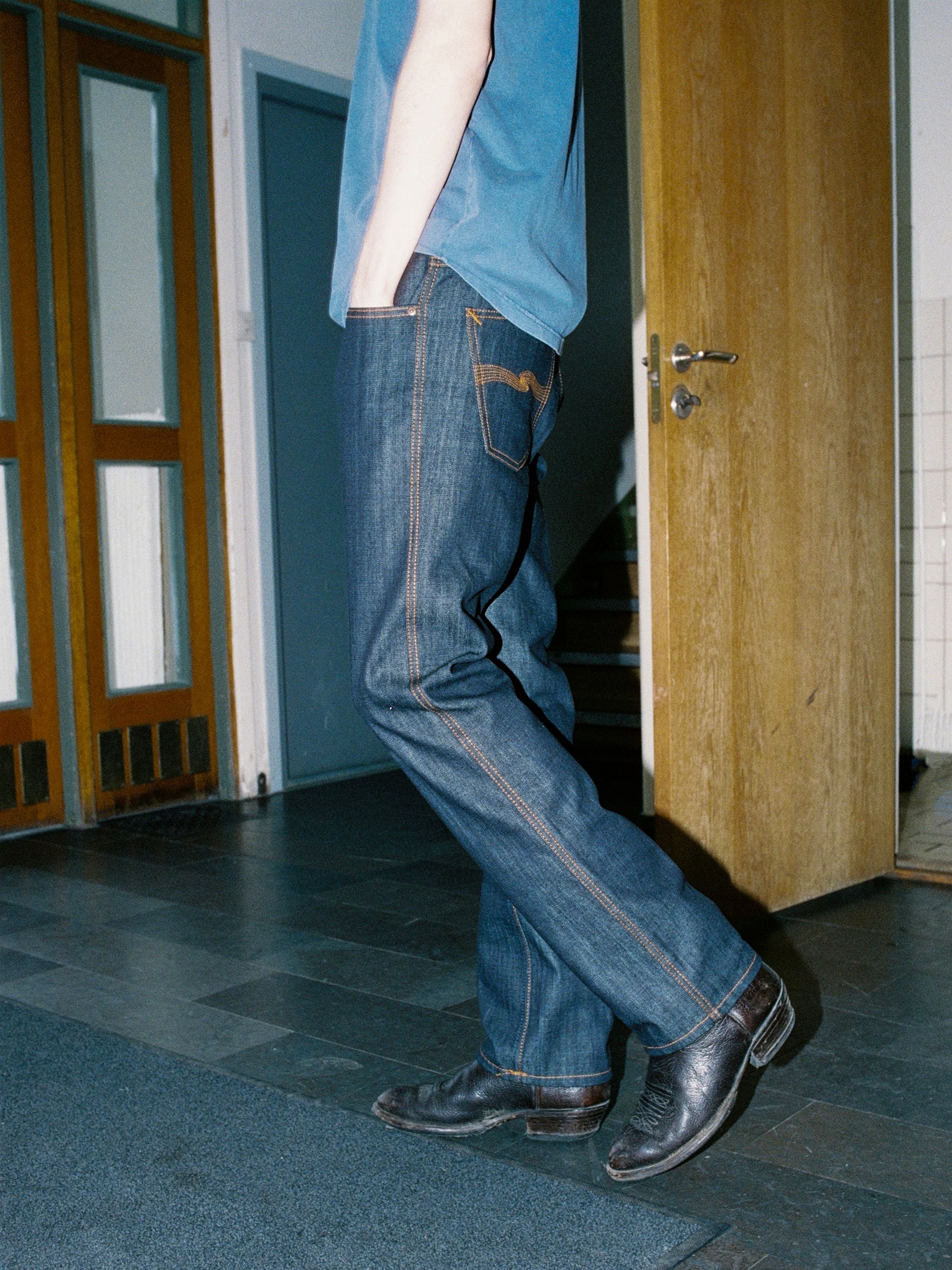 Nudie Jeans Slim Jim Dry Two, rigid indigo organic cotton slim straight jeans with orange stitching, worn by a model.