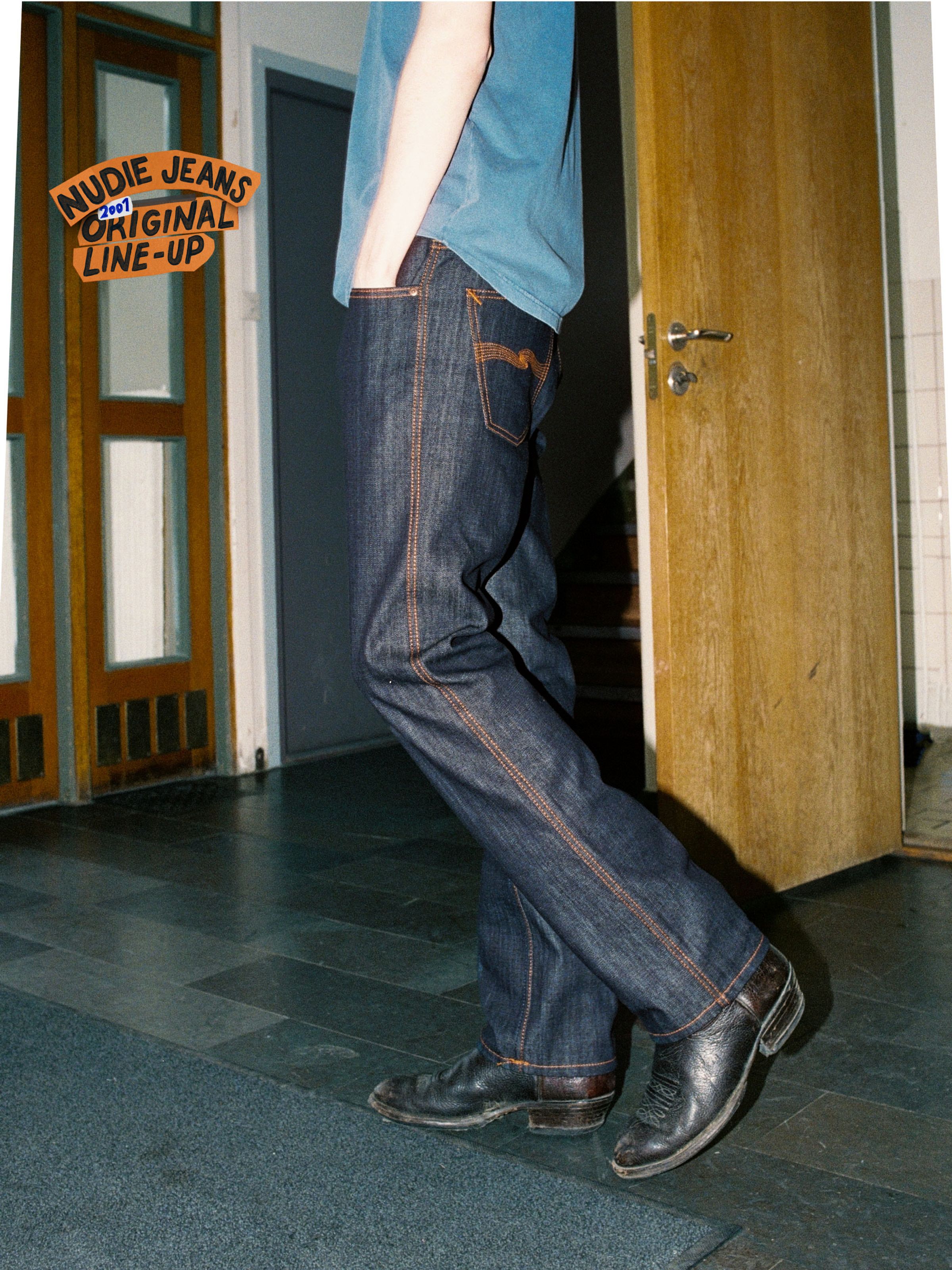 Nudie Jeans Slim Jim Dry Two, men's slim straight jeans in dry blue denim with orange seams, side view on a model.