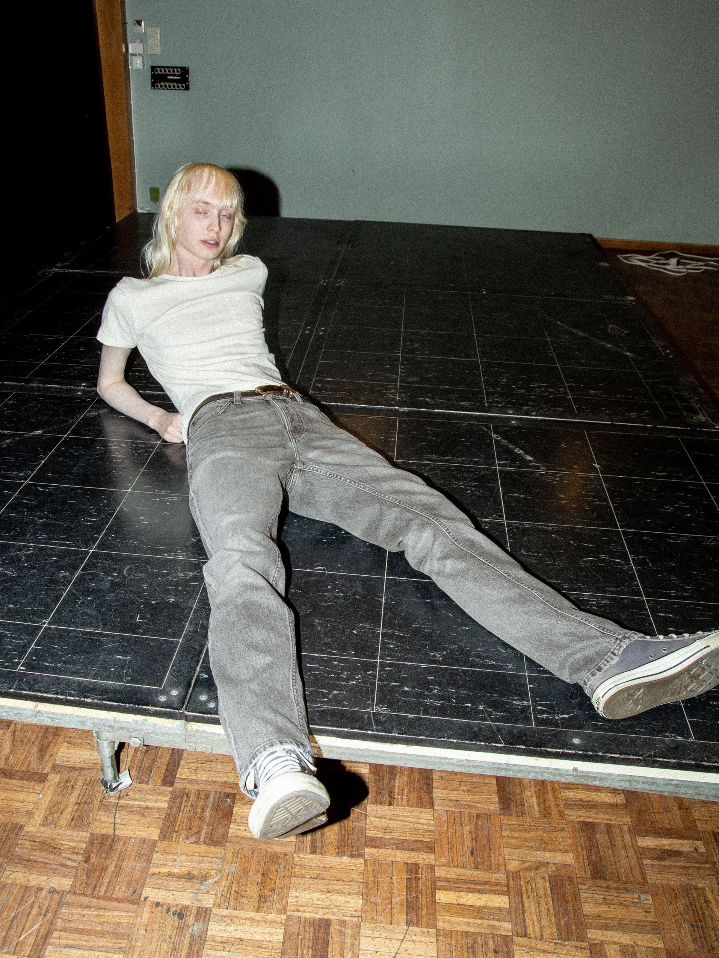 Nudie Jeans Solid Ollie Pale Ash, men's slim-fit grey wash jeans shown on a model reclining on a platform.