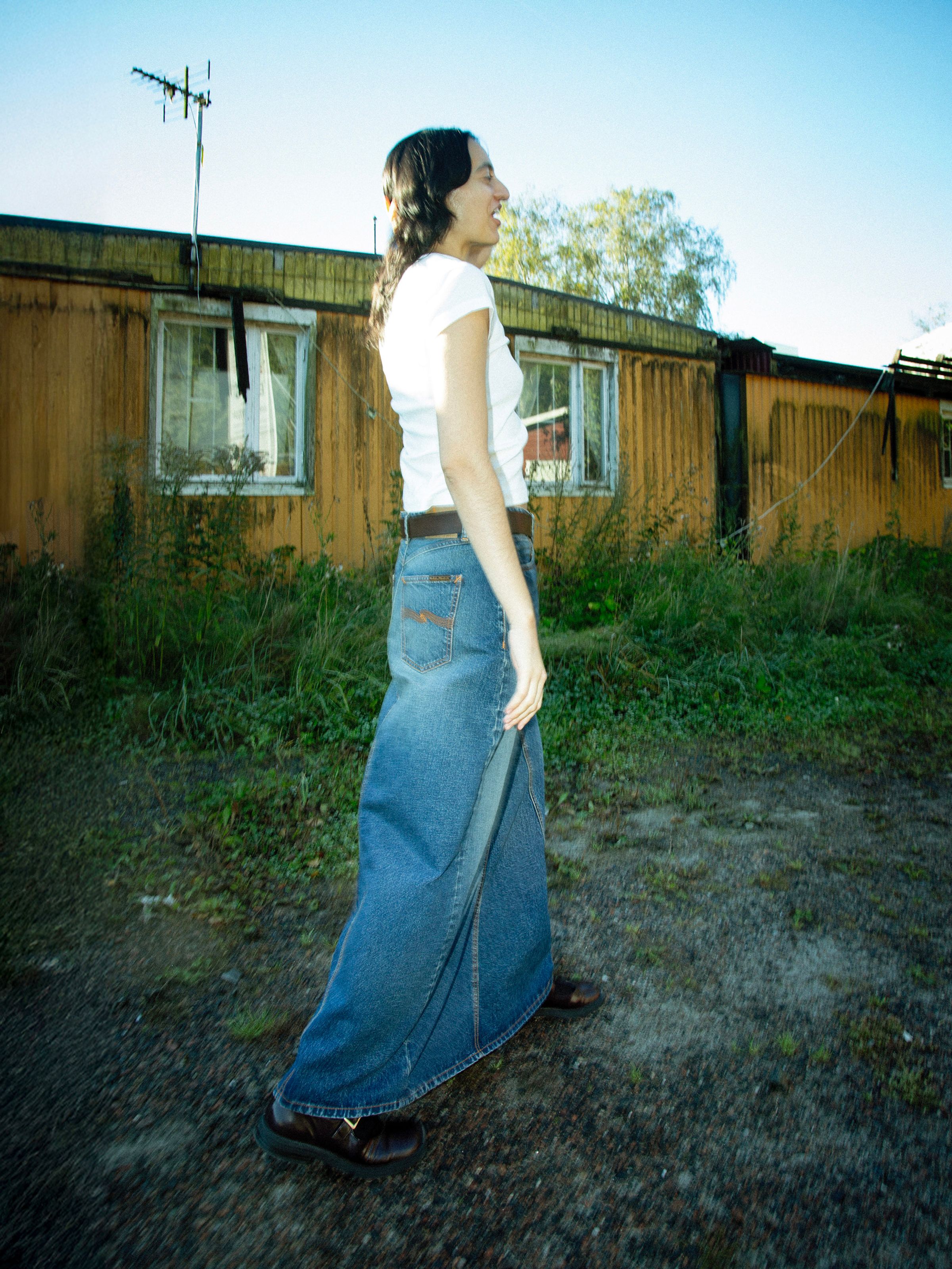 Nudie Jeans Tanja Skirt Orange Painter, a full-length denim skirt in a mid-blue wash, shown from the side on a model.