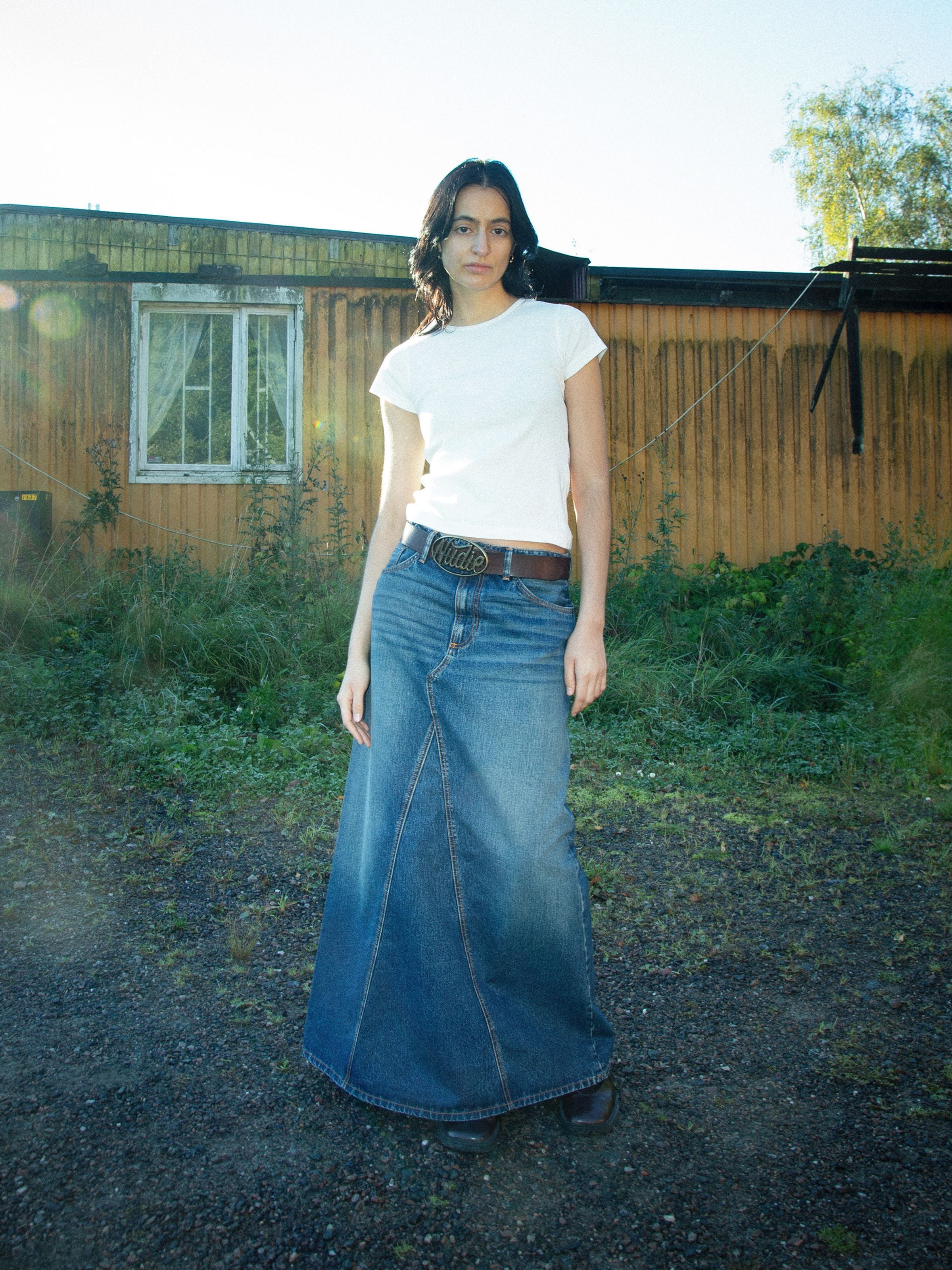 Nudie Jeans Tanja Skirt Orange Painter, ein bodenlanger Jeansrock an einem Model in einer Outdoor-Lifestyle-Umgebung.