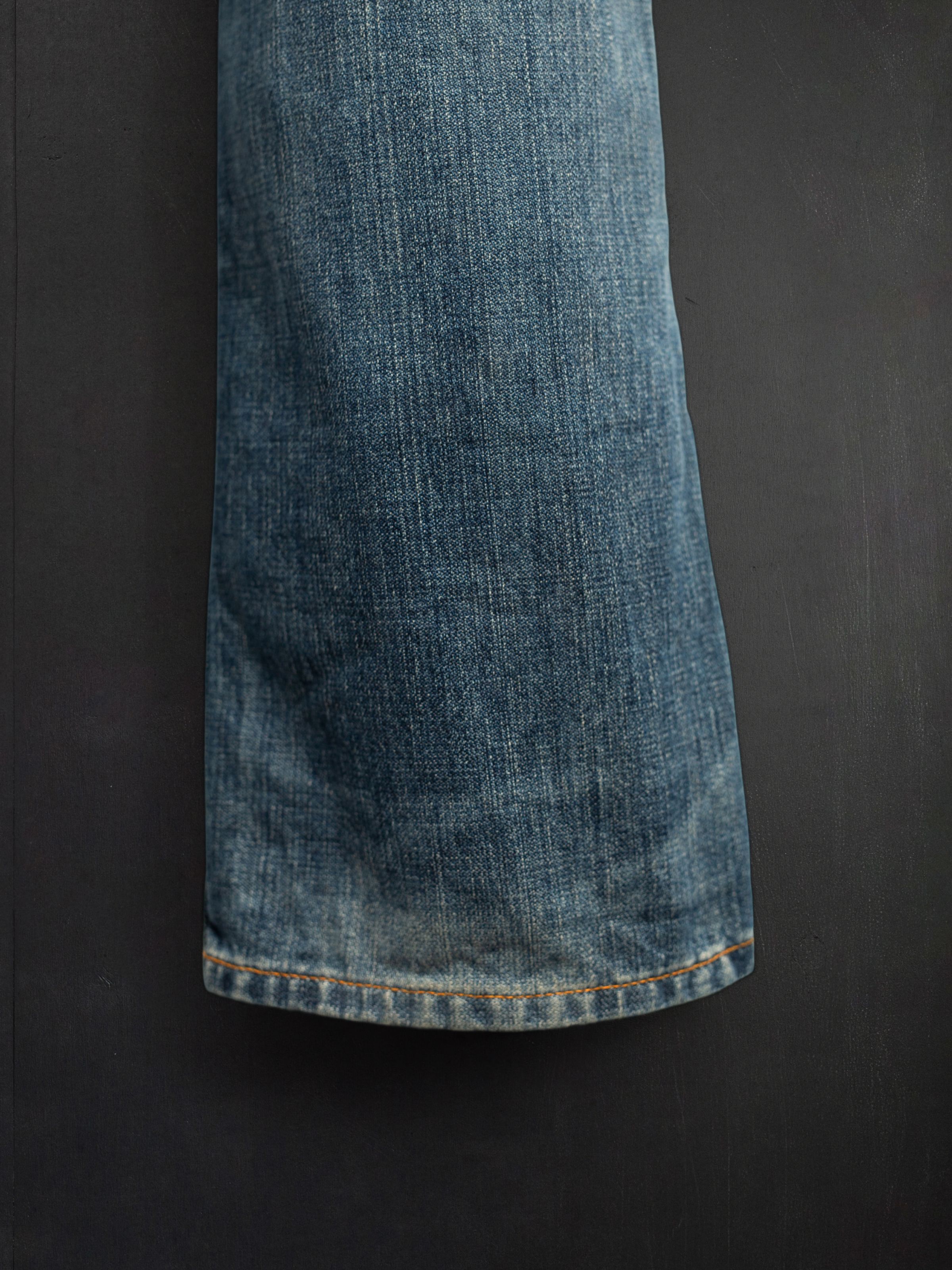 Nudie Jeans Slim Jim Quarter Tracks, a close-up of the leg hem in mid blue rigid denim with orange thread stitching.
