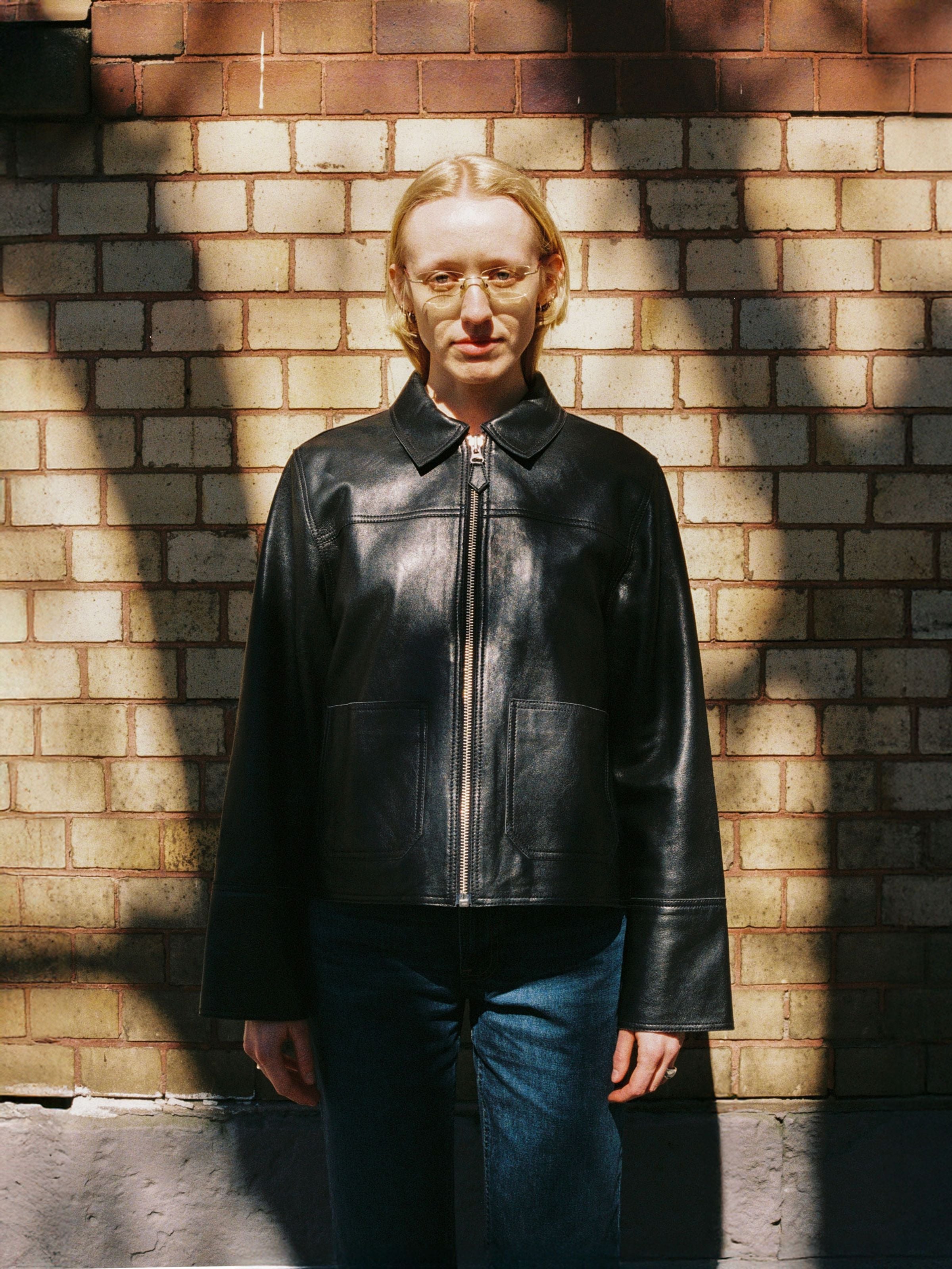 Nudie Jeans Anja Zip Jacket in black lamb skin leather, shown on a model against a brick wall background.