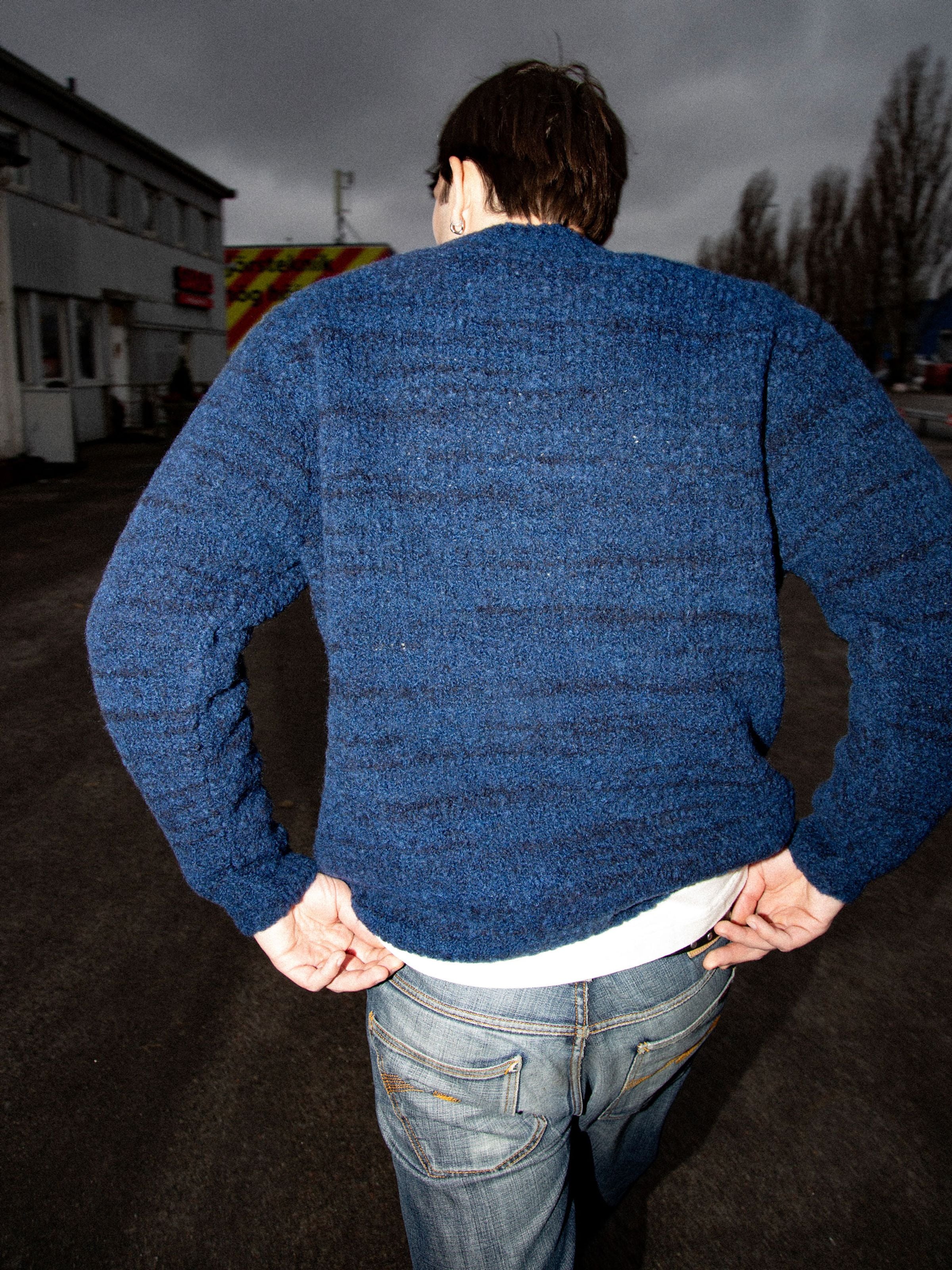 Nudie Jeans Gurra Boucle Sweater Multi, a textured blue organic wool blend sweater shown from the back on a model outdoors.