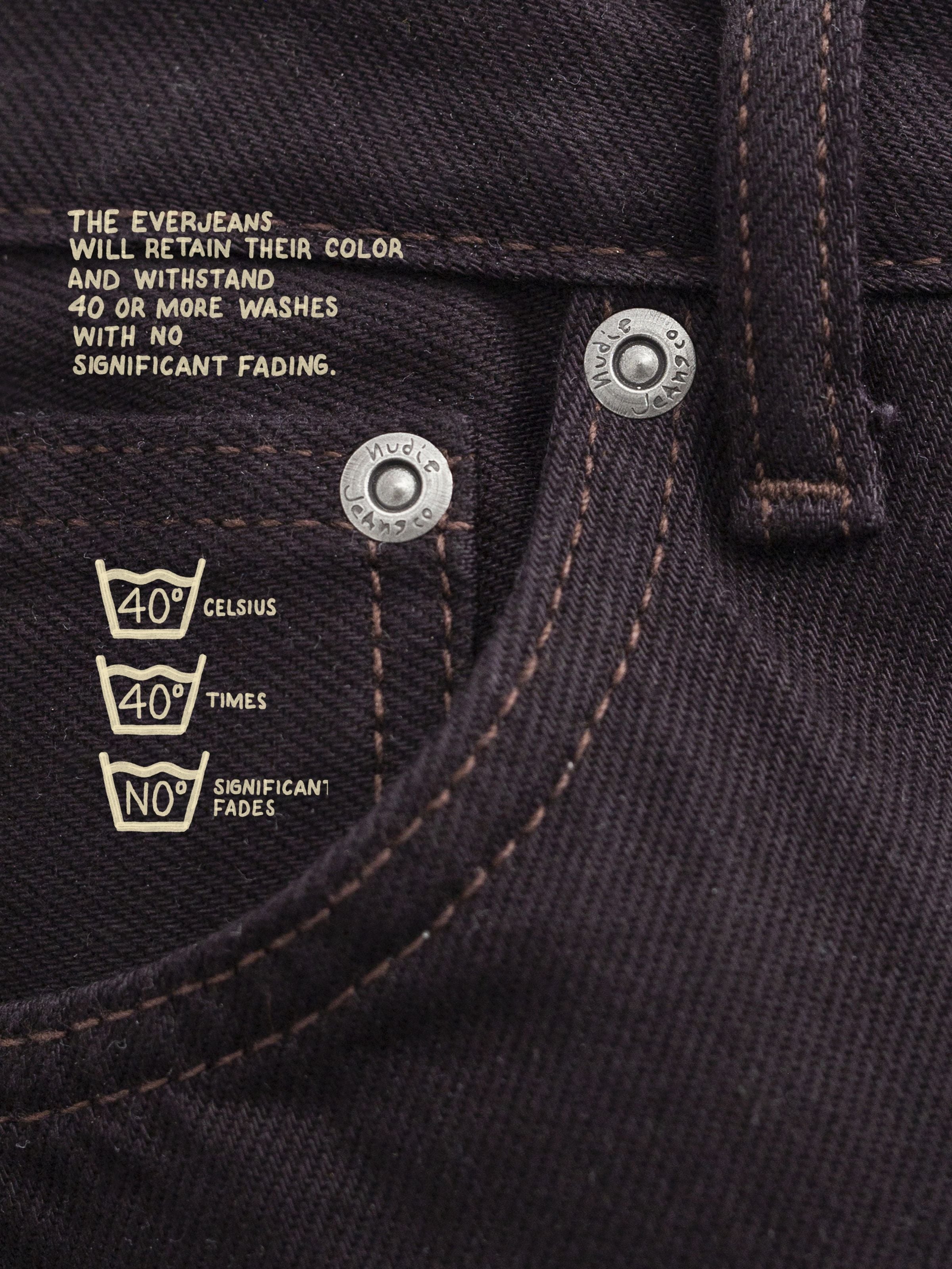 Nudie Jeans Rad Rufus Everbrown, Detailaufnahme von tiefbraunem Rigid Denim mit silbernen Nieten und aufgedrucktem Text.