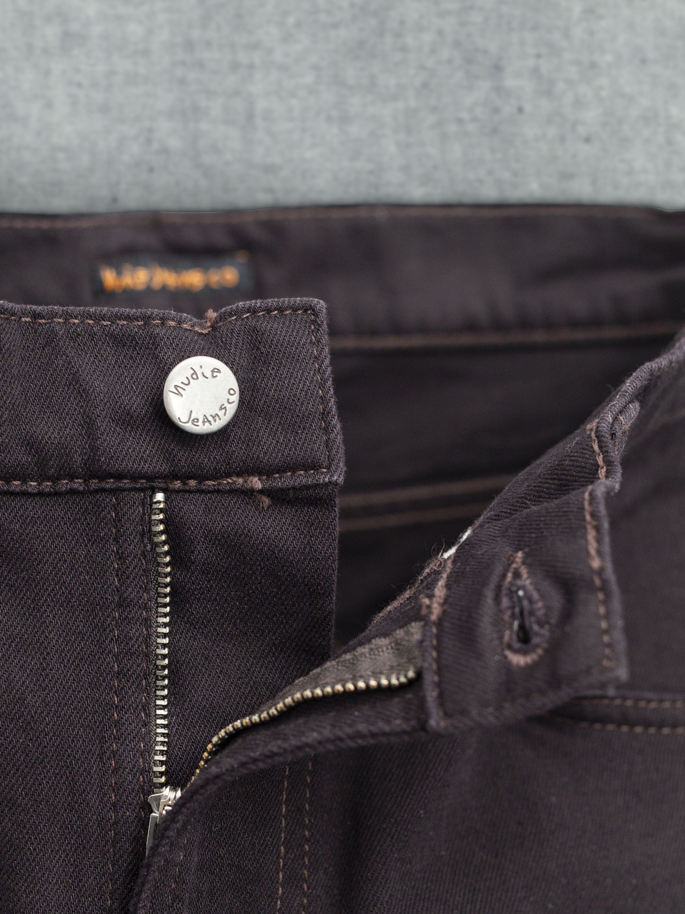Nudie Jeans Lean Dean Everbrown, Nahaufnahme des altsilbernen Knopfs und Zip Fly auf tiefbraunem Denim aus Bio-Baumwolle.