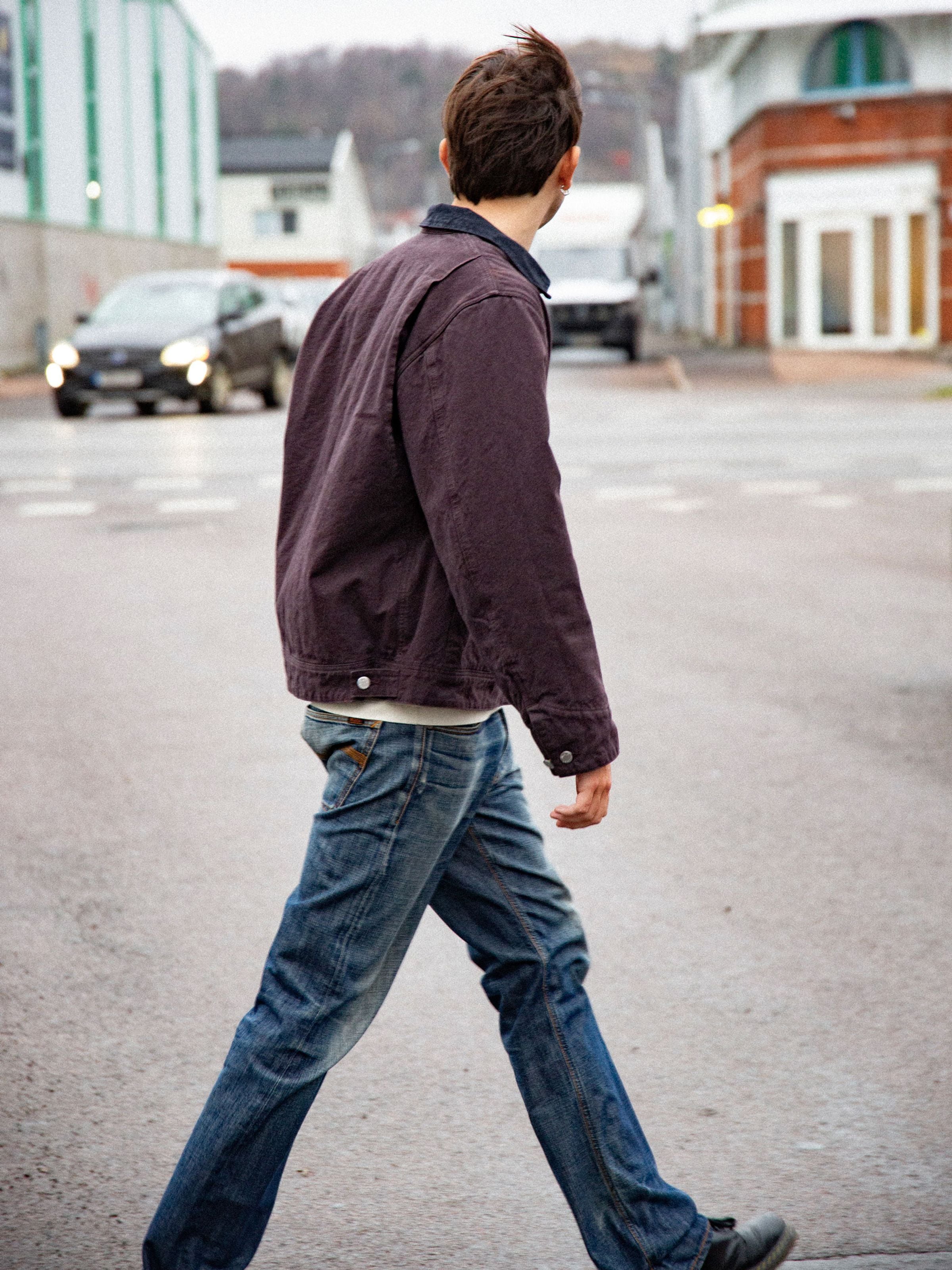 Nudie Jeans Blake Zip Jacket aus dunkelbraunem Bio-Baumwoll-Duck-Canvas, von hinten an einem Model im Freien gehend.
